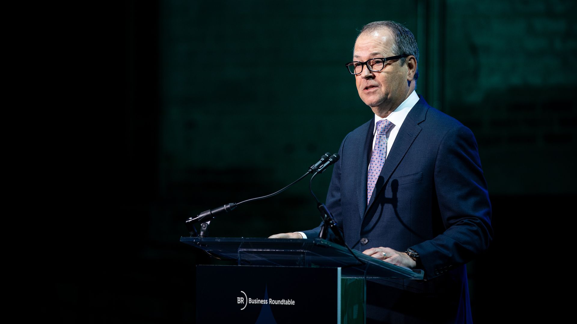 Man in a dark blue suit and patterned tie speaking at a podium with microphones, with a dark green background and a sign reading "Business Roundtable."