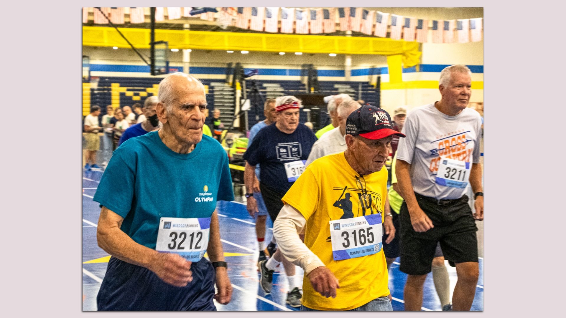 Indoor track meet with older runners wearing bibs 3212, 3165 and 3211. A teal-shirted man leads, followed by a yellow-shirted man and a white-shirted runner; crowd and bleachers visible in the gym.