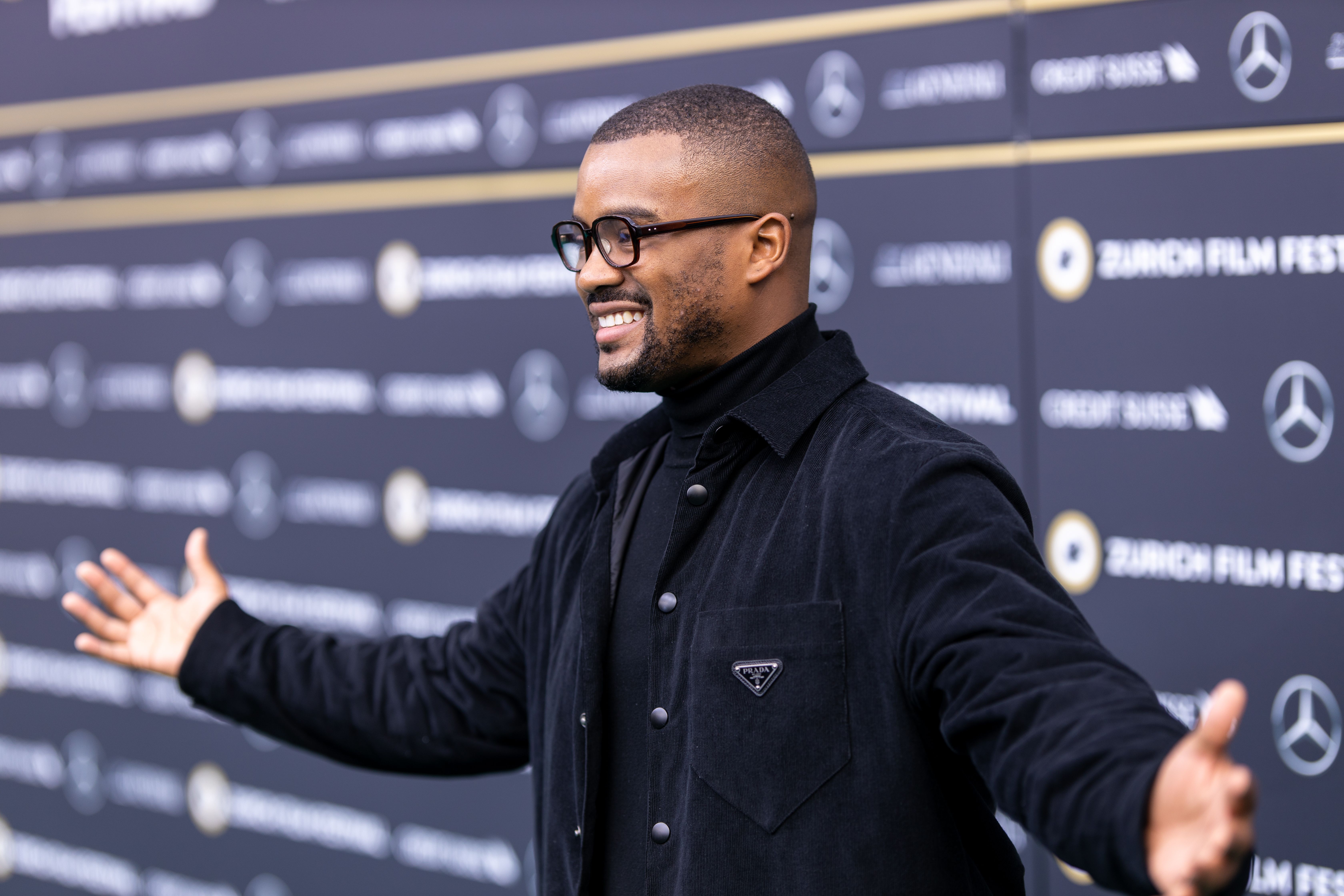 A man stands on a red carpet with his arms outstretched and smiling.