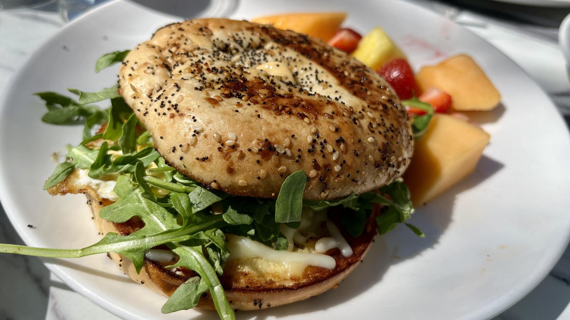 A sesame- and poppy-seed bagel with cream cheese and arugula on a white plate, plus fruit chunks (pineapple, melon, strawberries); bright sunny outdoor dining scene.