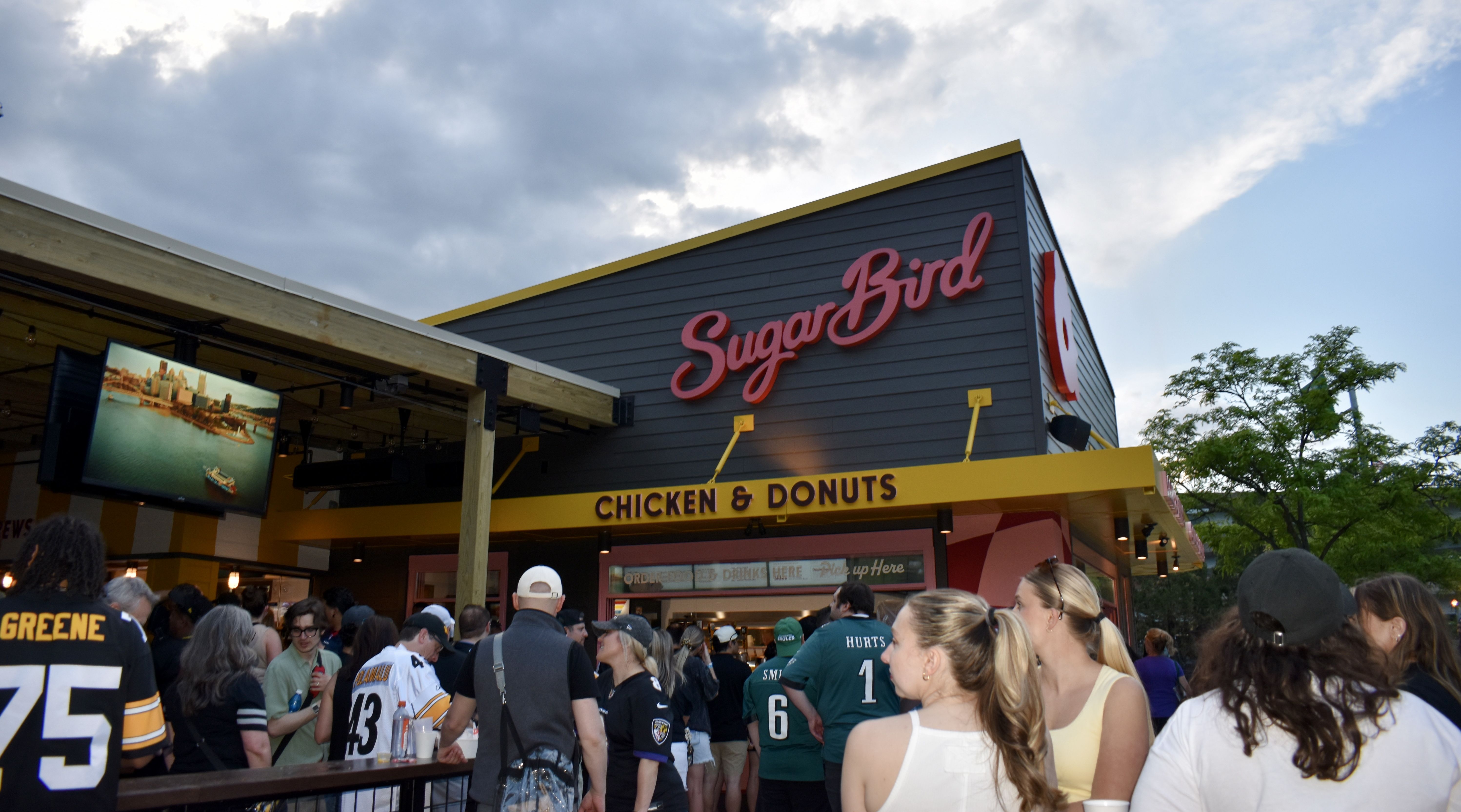 Busy outdoor scene in front of a gray building with a large red SugarBird sign and a yellow 'CHICKEN & DONUTS' banner. A TV screen shows a harbor, with a cloudy sky and trees in the background.