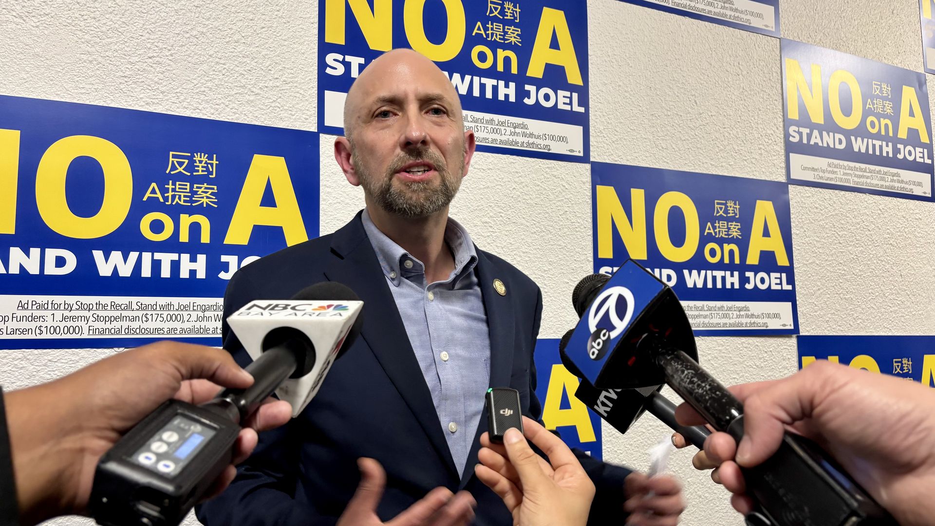A bald man in a dark suit speaks to reporters holding microphones labeled NBC Bay Area and abc7, in front of multiple blue and yellow posters reading "NO on A STAND WITH JOEL".