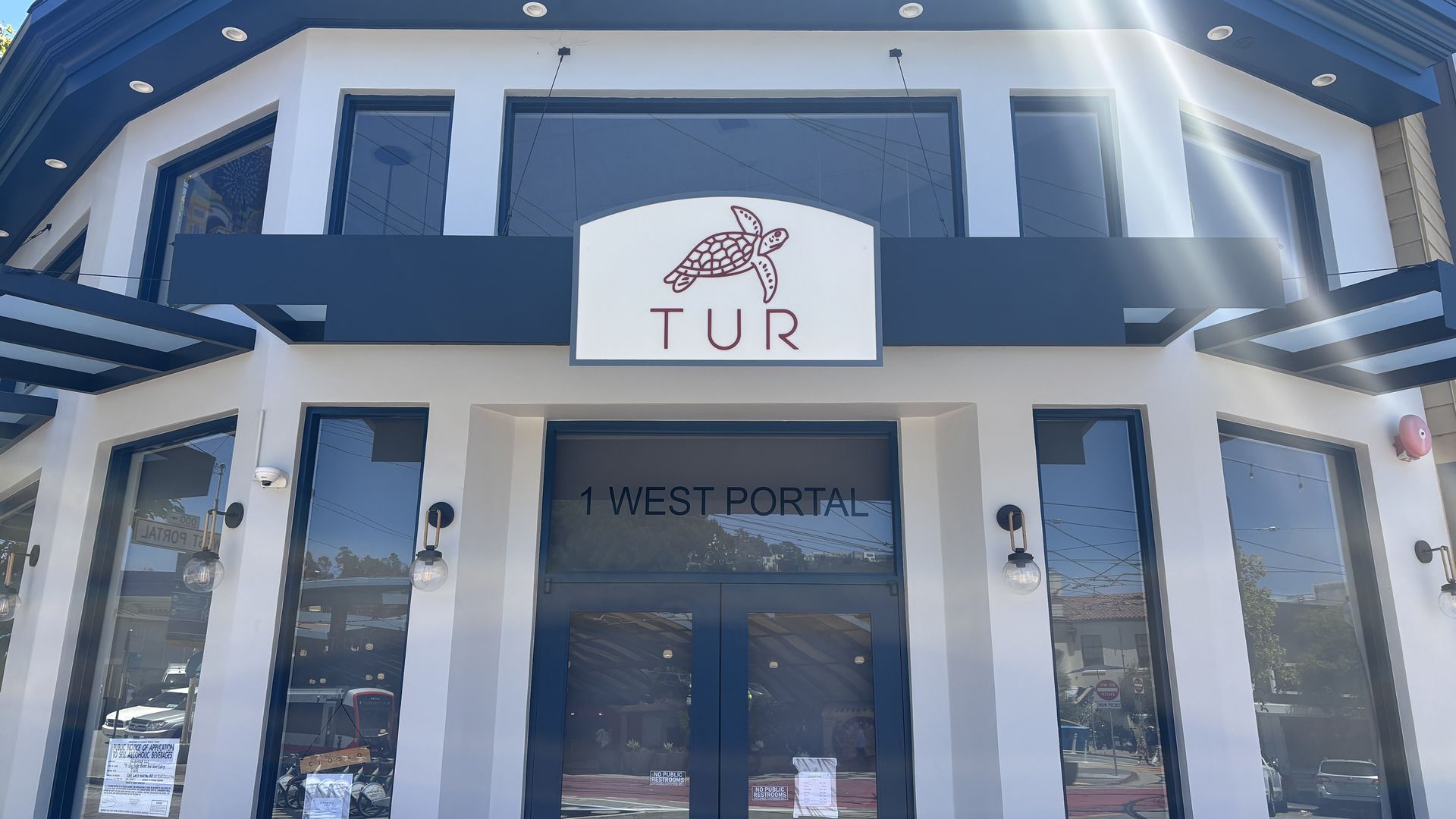 Modern white storefront with blue trim and awnings. A white sign with a red turtle drawing and the letters TUR sits above the entrance; large glass doors and windows reflect the sunny street.