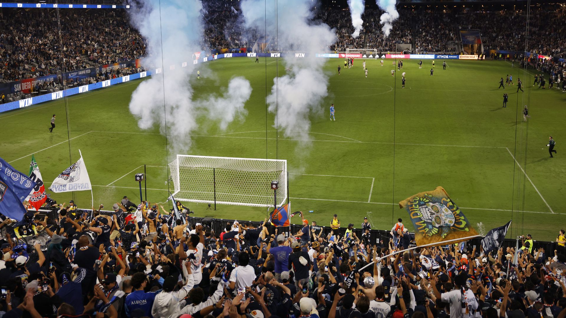 Fans cheer in the stands, waving big flags, as blue smoke and fireworks go off after San Diego FC wins soccer game at Snapdragon Stadium. 