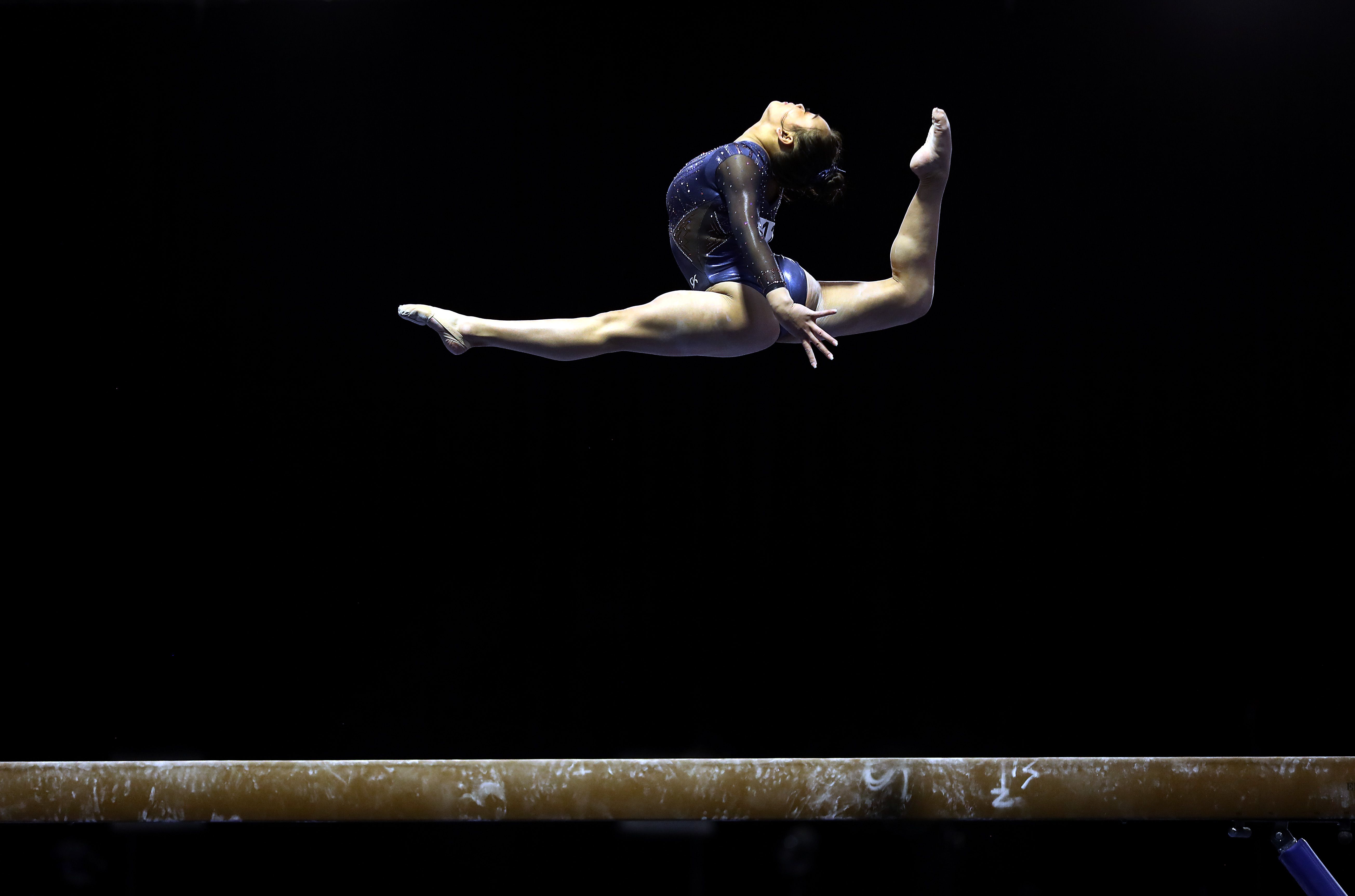 Photo of Suni Lee mid-split and raising her head to her back leg in the air over a beam