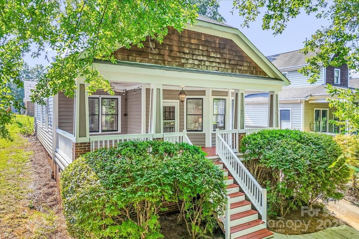 Gray house with white porch railings, stairs, and trimmed bushes in front, under bright daylight and surrounded by trees.