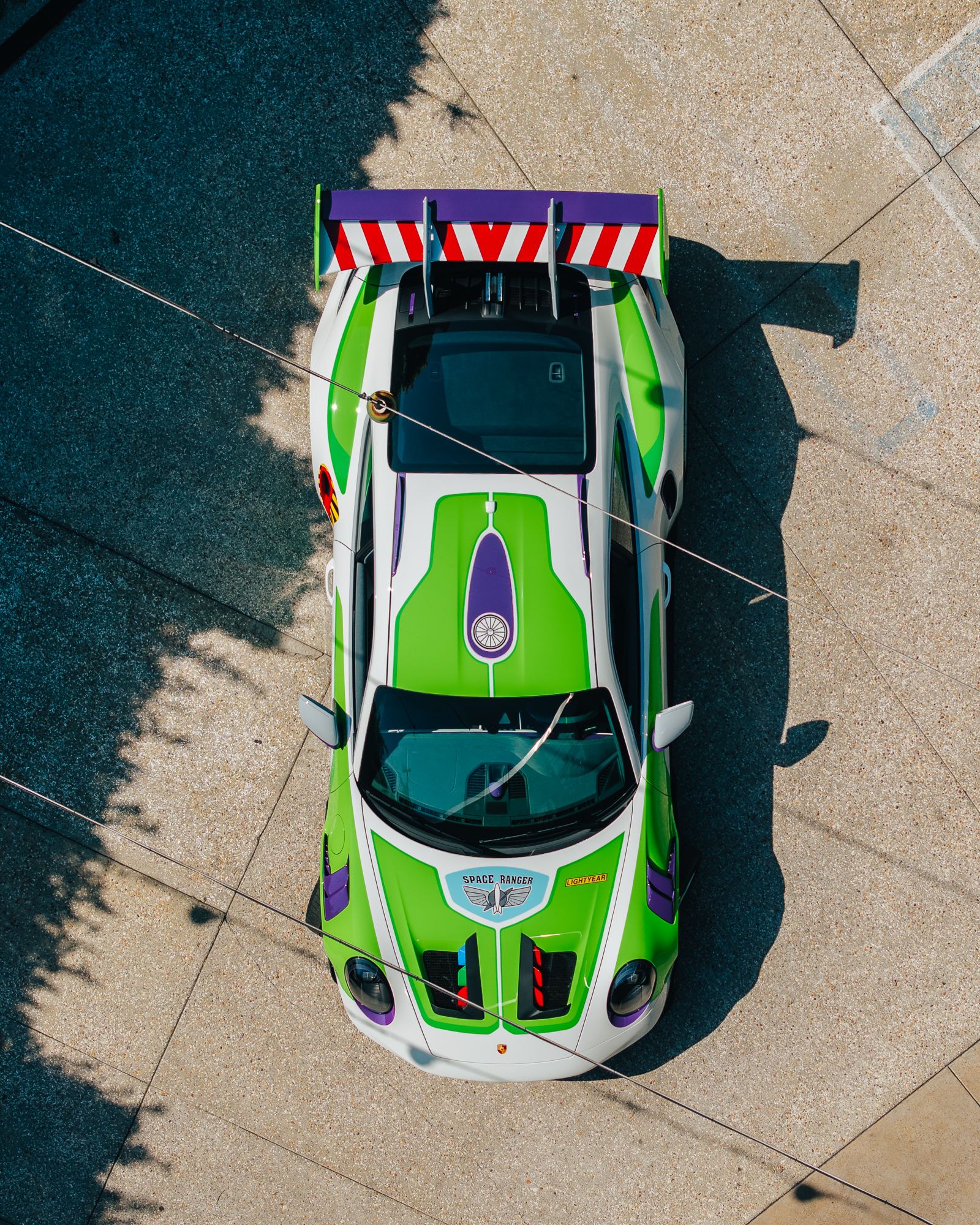 Top-down view of a neon green and purple race car with a red-and-white striped rear wing, parked on a sunlit concrete surface; diagonal power lines and tree shadows cross the scene.