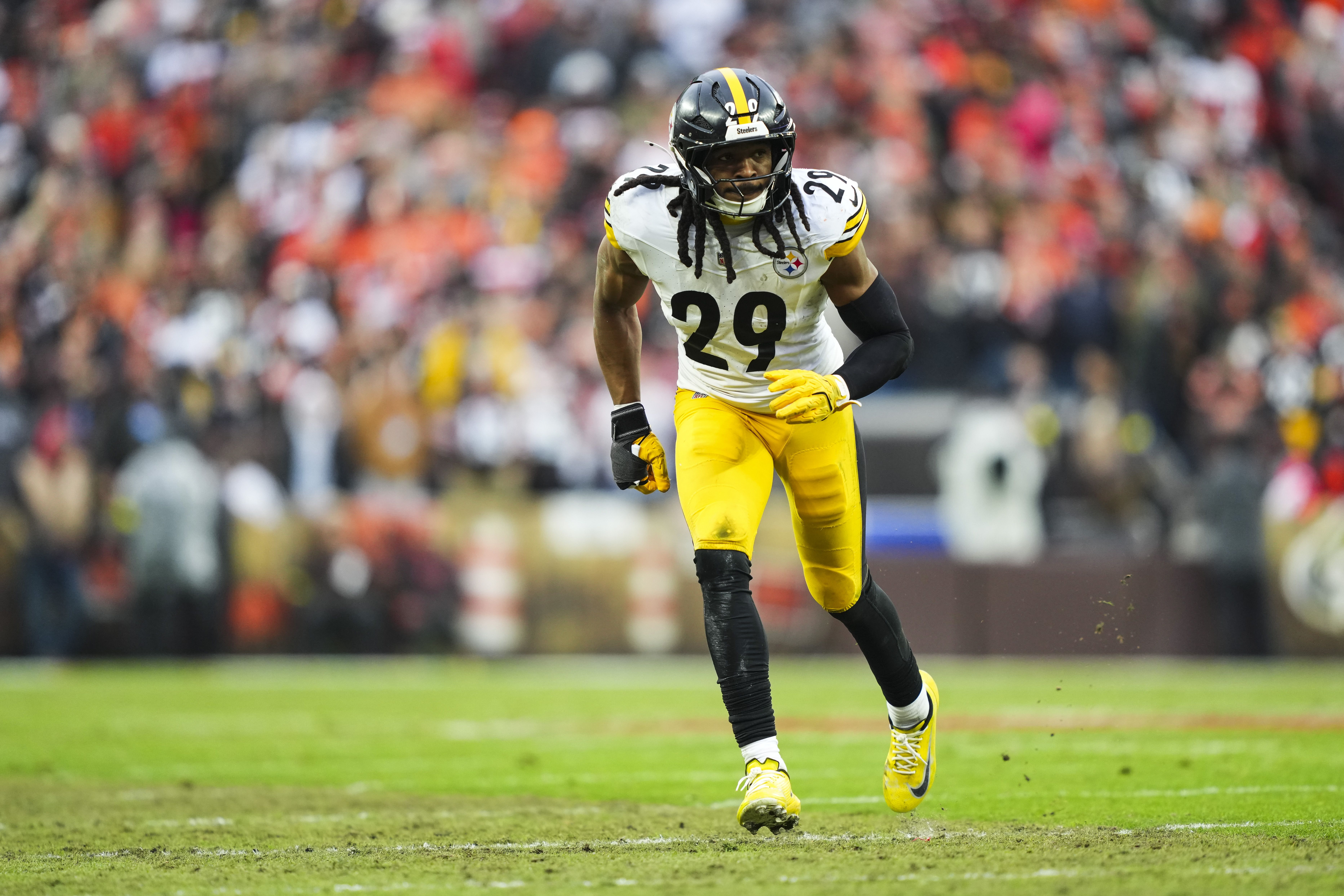 Football player wearing black helmet and yellow pants with number 29 on white jersey running on grassy field with blurred crowd in the background.