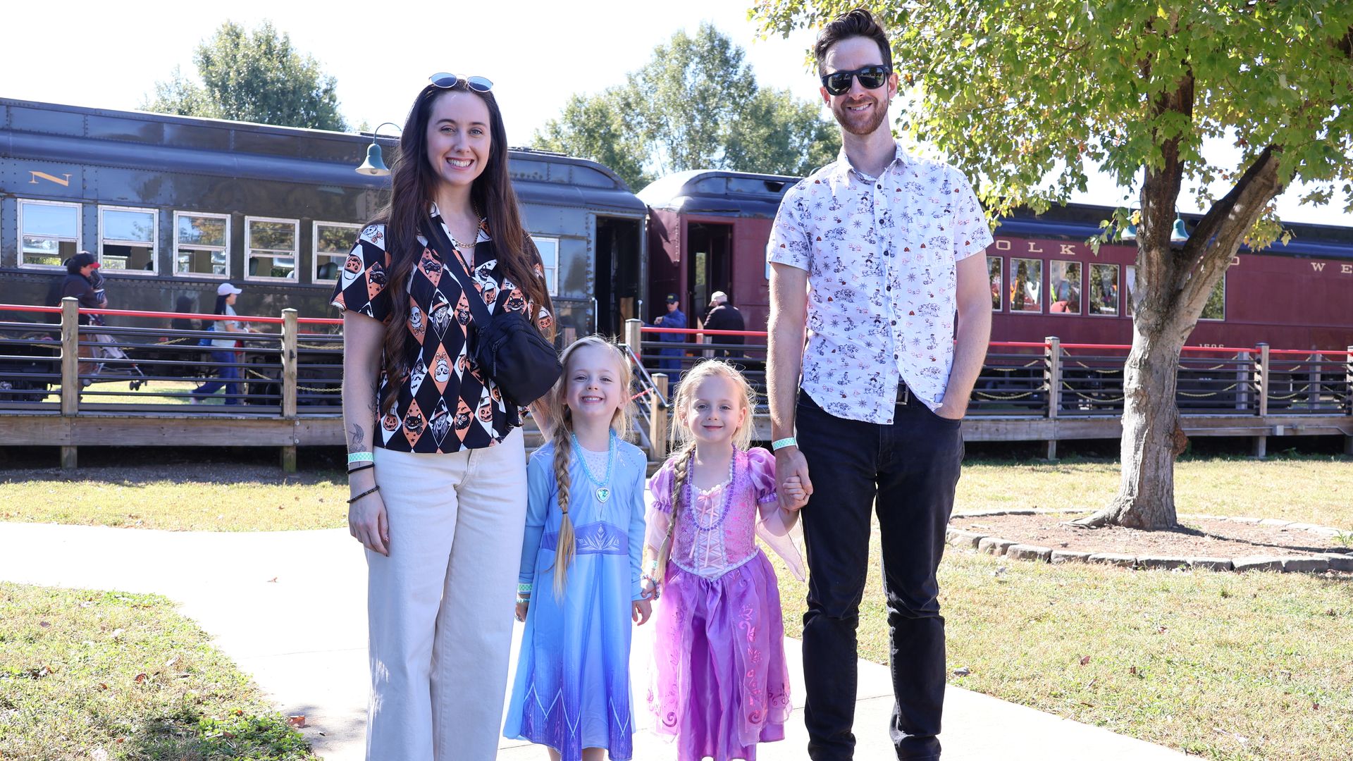 Family of four posing outdoors near a vintage train. Mom wears white pants and a patterned top, dad wears black jeans and a white shirt. Two girls in princess dresses stand between them.