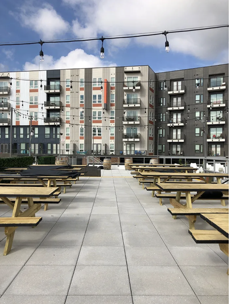 Outdoor courtyard with string lights and rows of wooden picnic tables on a tiled surface, with a modern gray, white, and red apartment building in the background under a blue sky.