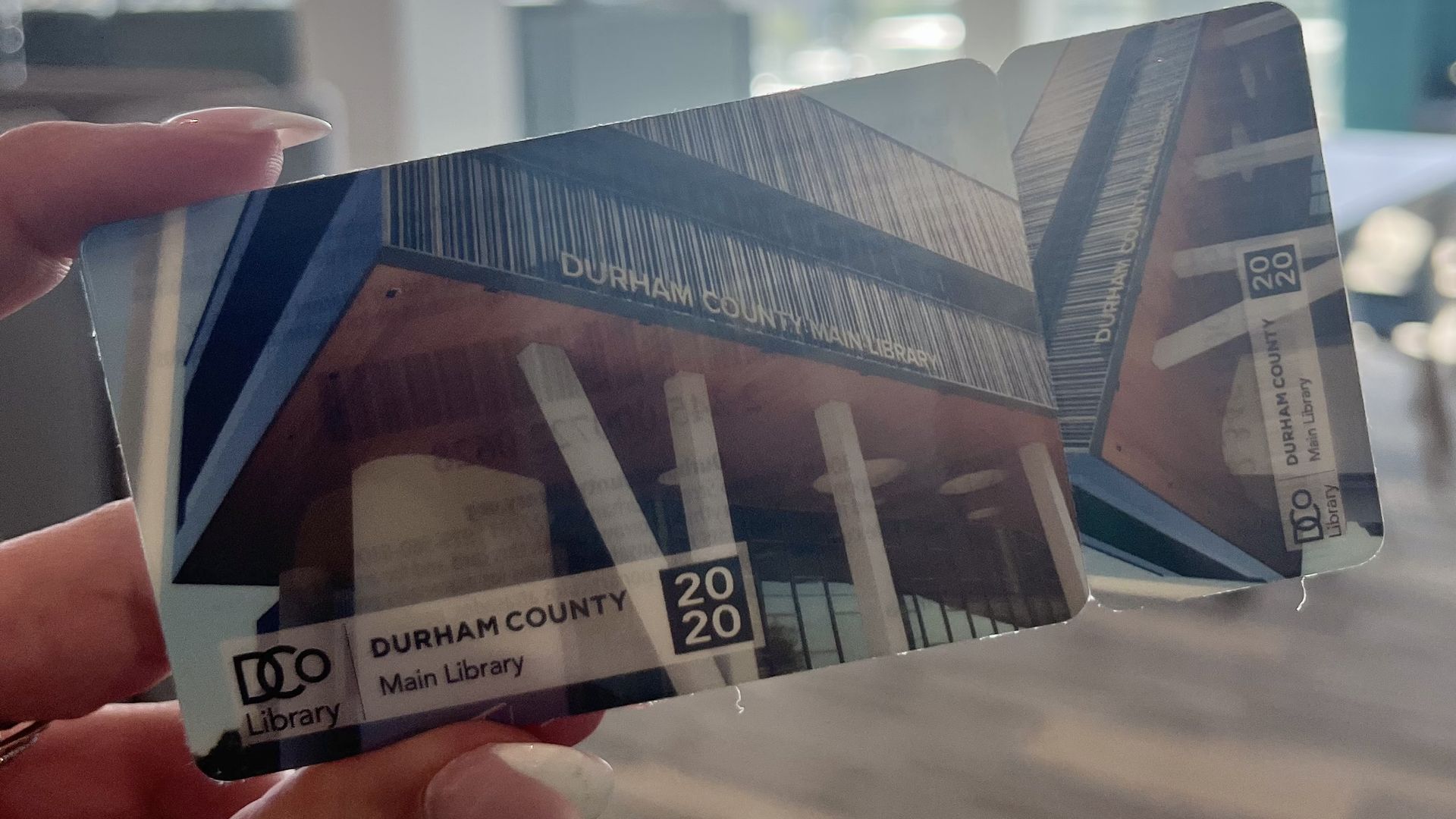 Close-up of a hand holding a Durham County Library Main Library card; blurred background of a bright library with large windows and curved ceiling lights.
