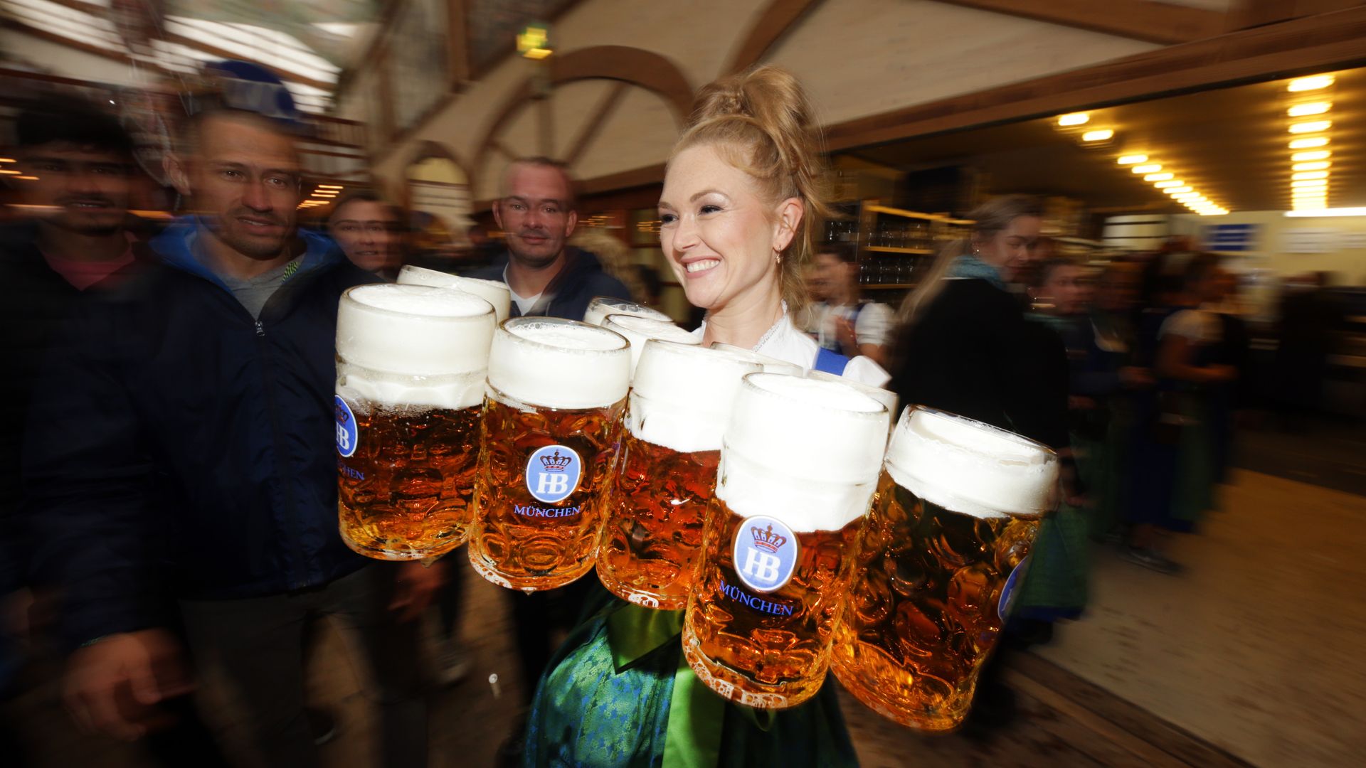 Smiling woman in a dirndl carries several large frothy beer steins labeled HB München at a crowded festival hall, with motion-blurred crowd and warm lights in the background.