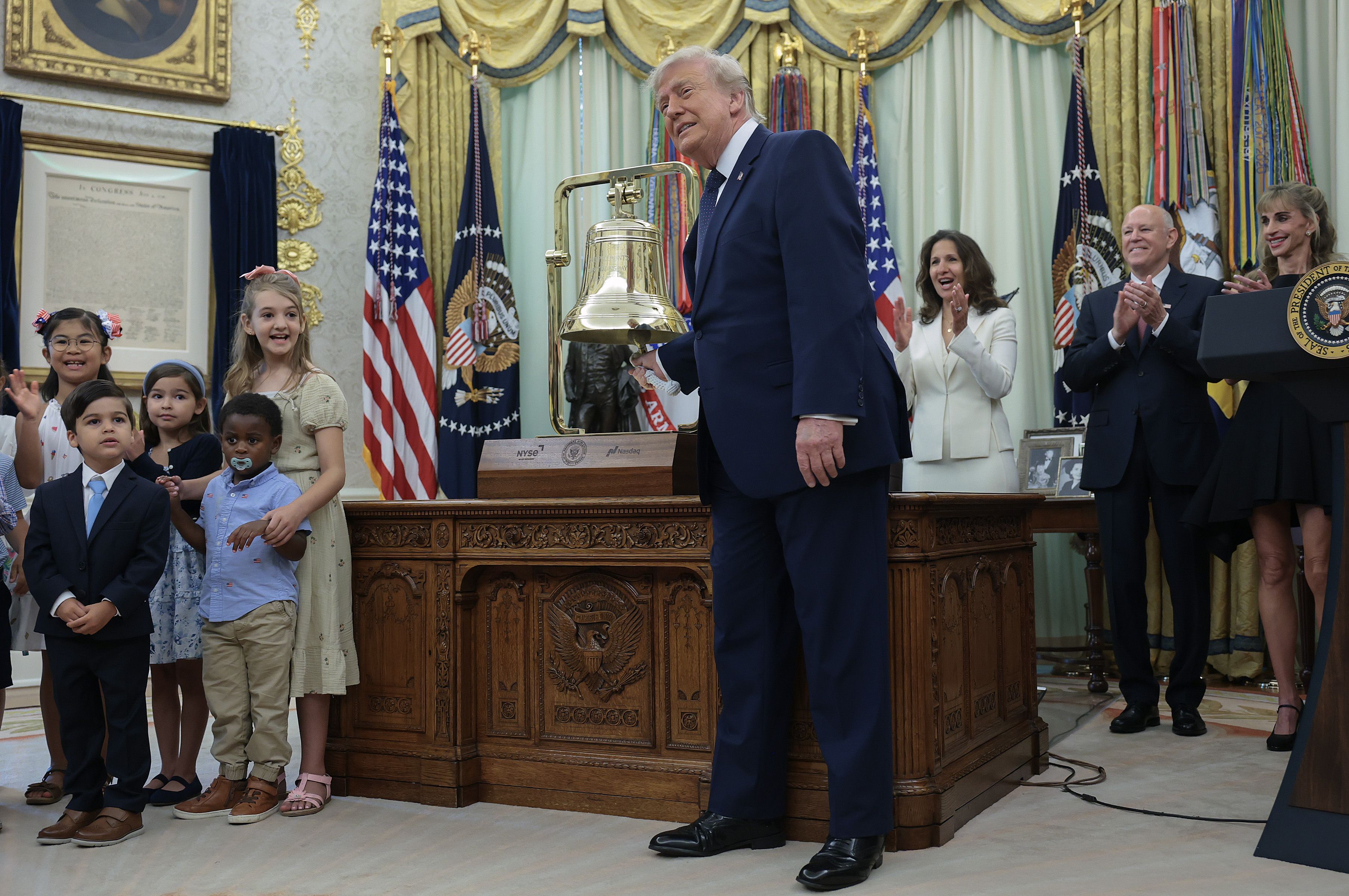 President Trump rings a golden bell on his desk while people clap behind him and a group of children stand beside him.