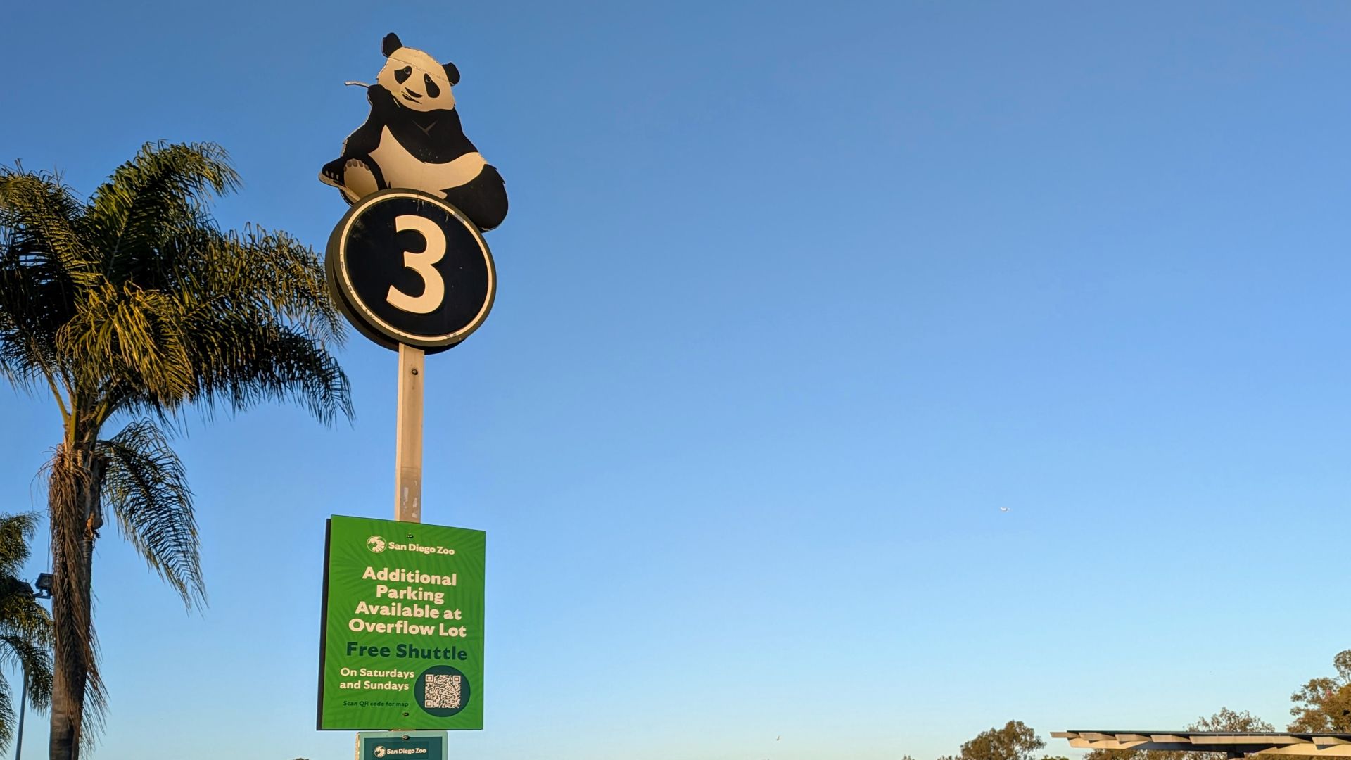 Parking lot at San Diego Zoo with a sign featuring a panda above number 3, green sign for extra parking and shuttle, cars parked under clear blue sky near palm trees.