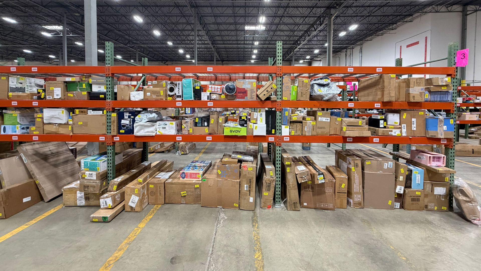 Wide warehouse aisle with tall orange metal racks filled with cardboard boxes and packing, fluorescent lights overhead, concrete floor, gray ceiling, and scattered pallets.