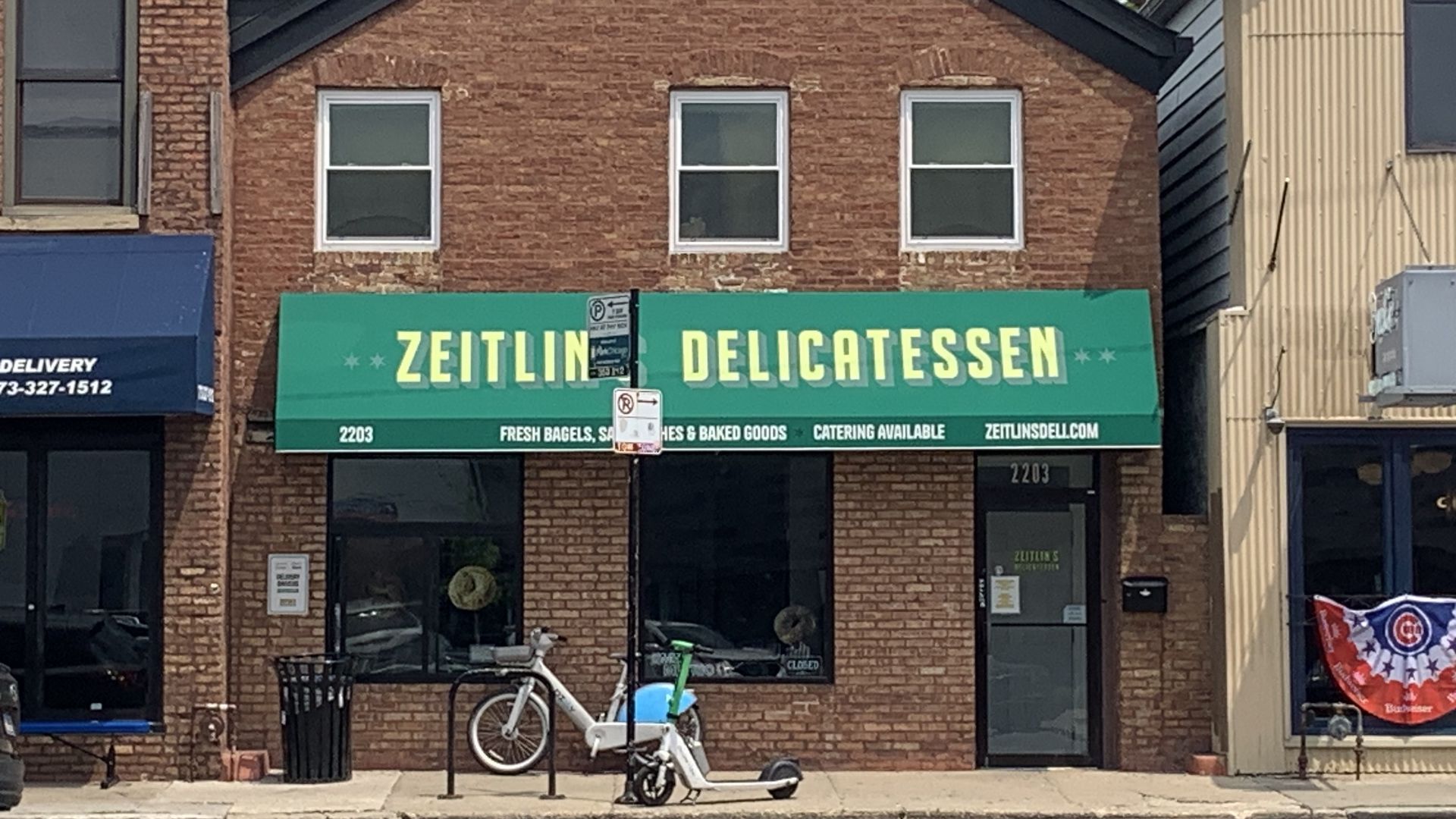 Brick storefront with a bright green sign reading "ZEITLIN DELICATESSEN" above three white-framed windows; bicycles parked out front, blue awning to the left, beige building to the right.