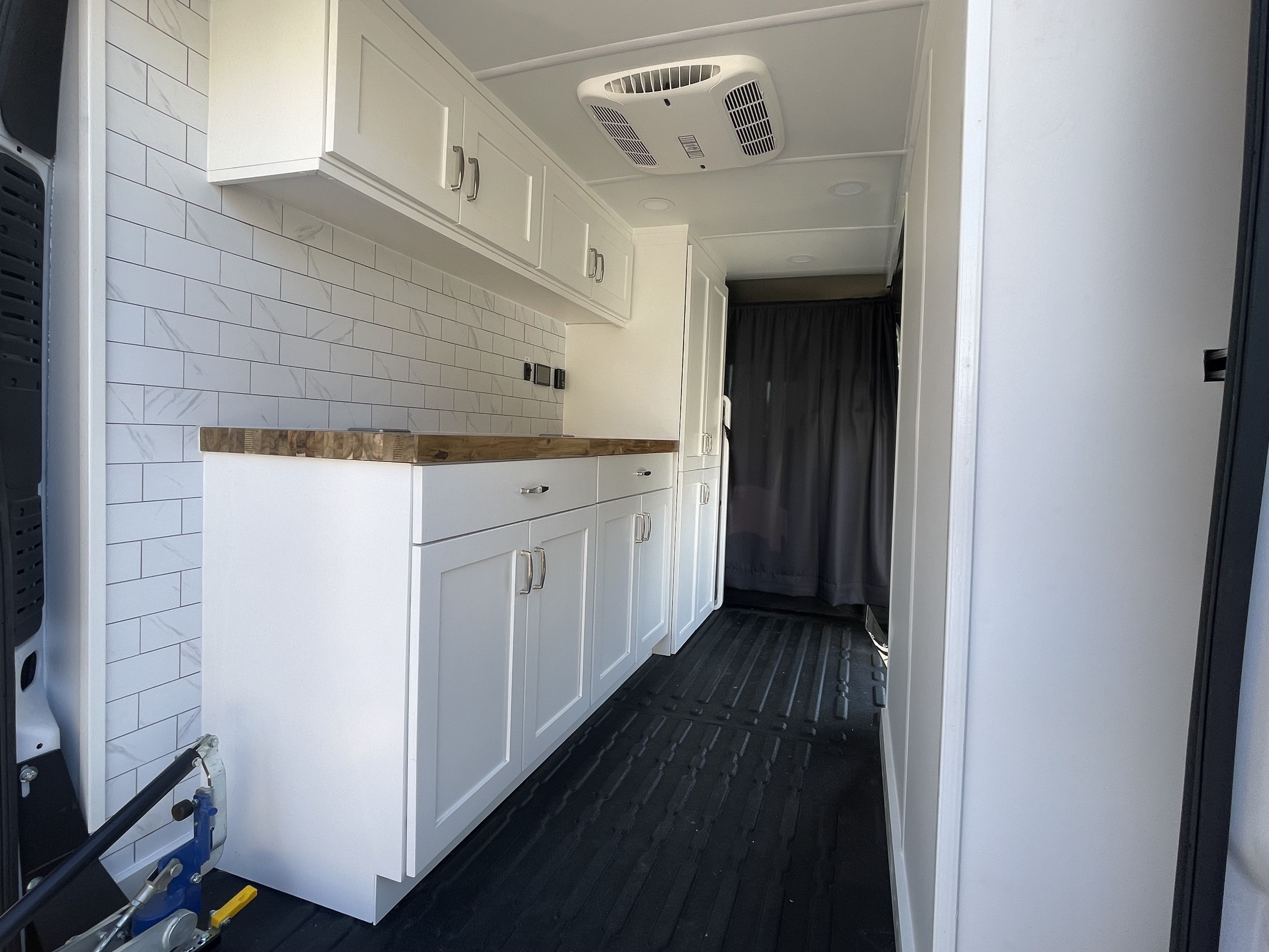 Interior of a compact white kitchen with white cabinets, wood countertop, white tile backsplash, black ribbed floor, ceiling vent, and a dark curtain at the end of the space.