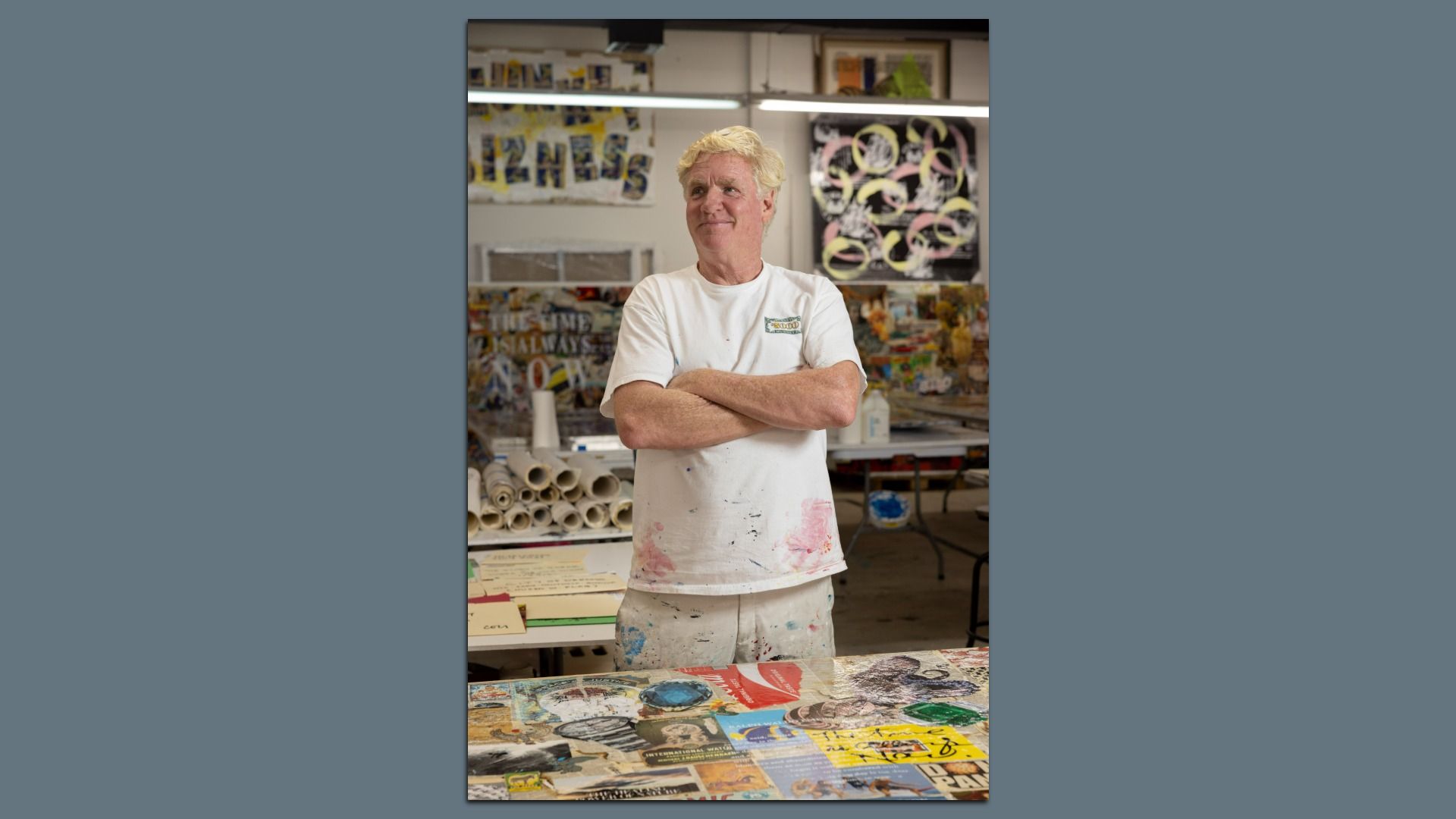 A man with blonde hair wearing a paint-stained white shirt and pants stands with arms crossed in an art studio filled with colorful posters and rolled-up canvases.