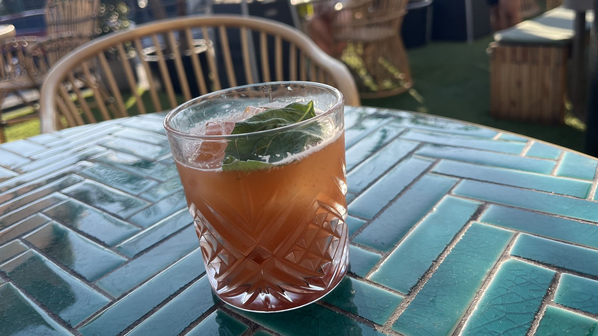A cocktail glass filled with an orange liquid topped with a basil leaf sits on a teal tiled table on an outdoor patio.