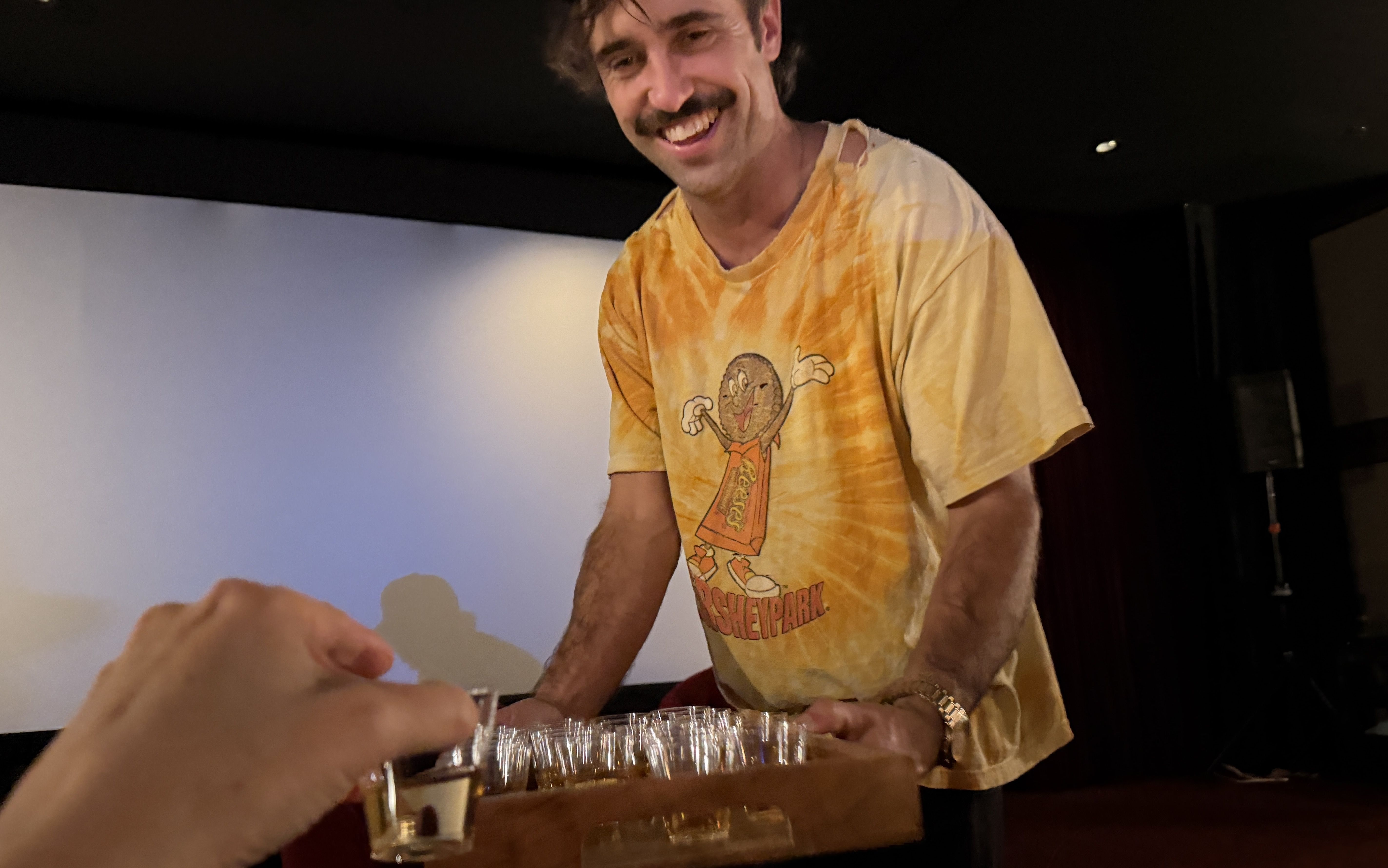 Man with mustache wearing a yellow tie-dye Reese's shirt serving tray of shot glasses to someone holding a shot glass in a dimly lit room with dark walls.