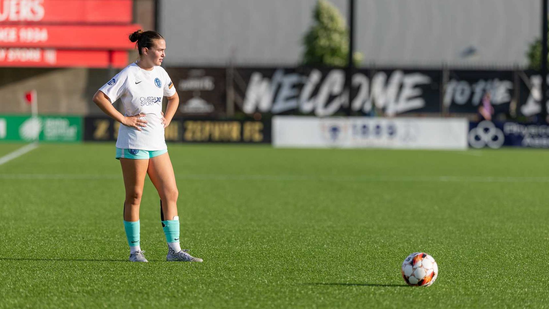 Female soccer player in white jersey and teal shorts standing on green field with hands on hips, looking at a ball on the grass during a sunny day at a stadium.