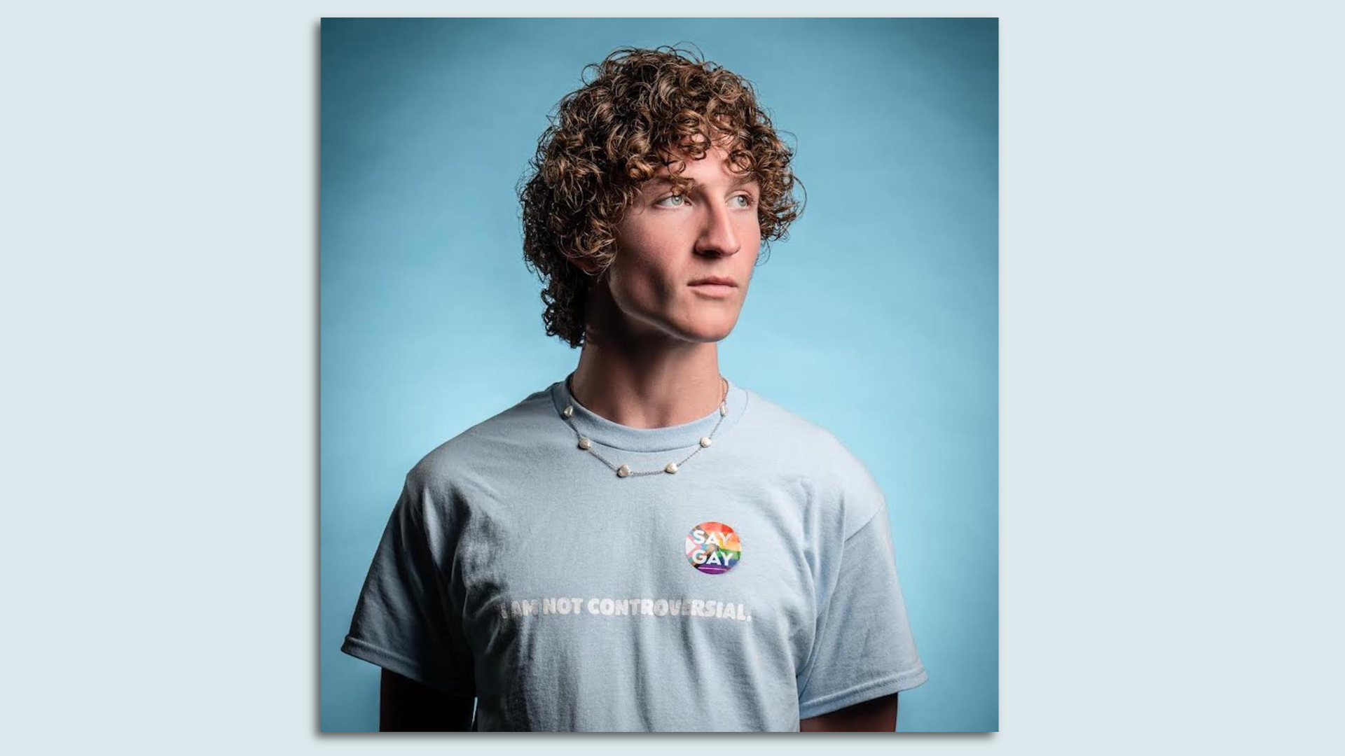 A headshot of Zander Moricz wearing a light blue shirt and a rainbow sticker, staring off into the distance.