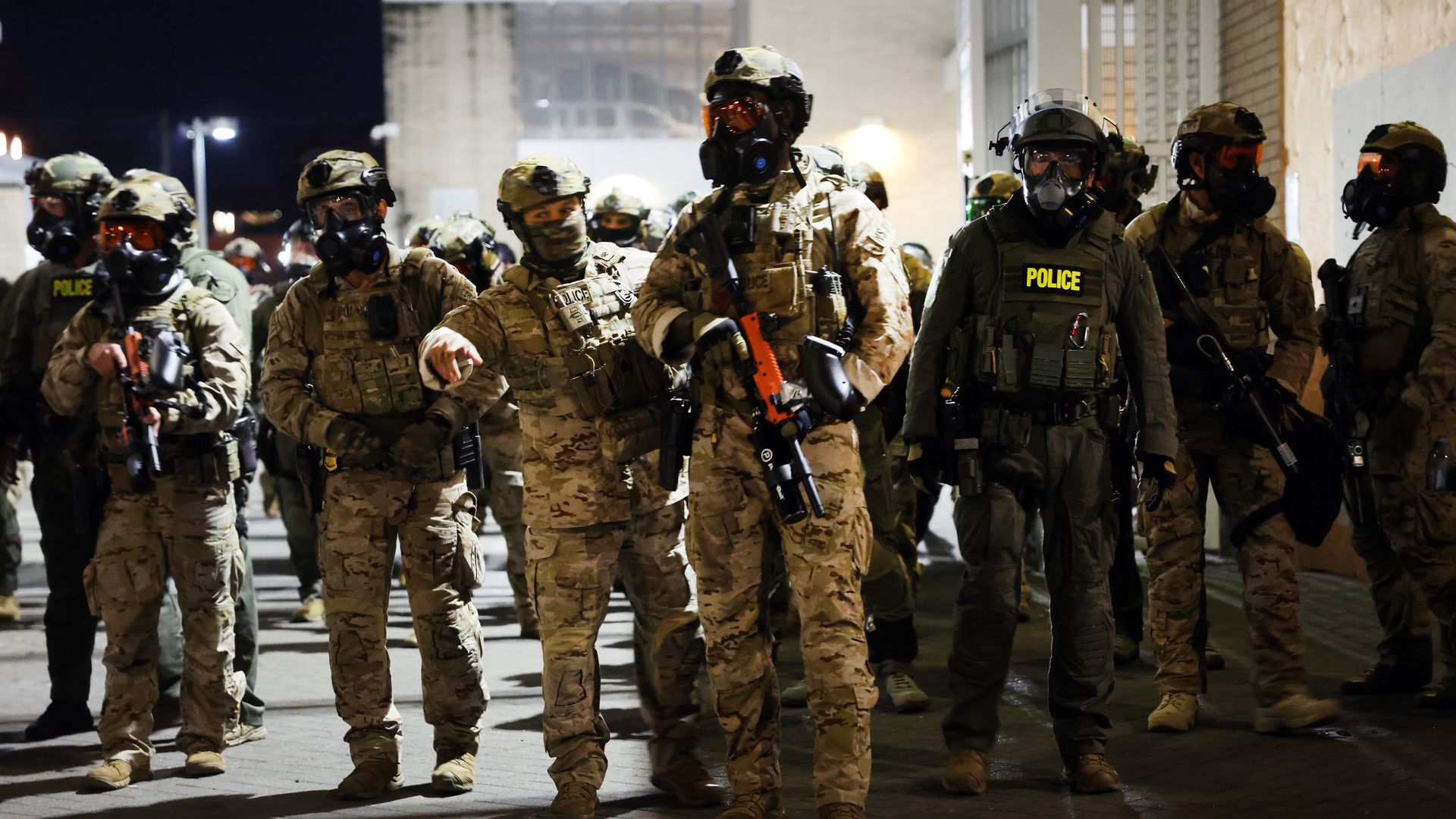 Group of police officers in tactical gear and gas masks standing outside at night, some in camouflage uniforms and others in dark green, with rifles in hand.