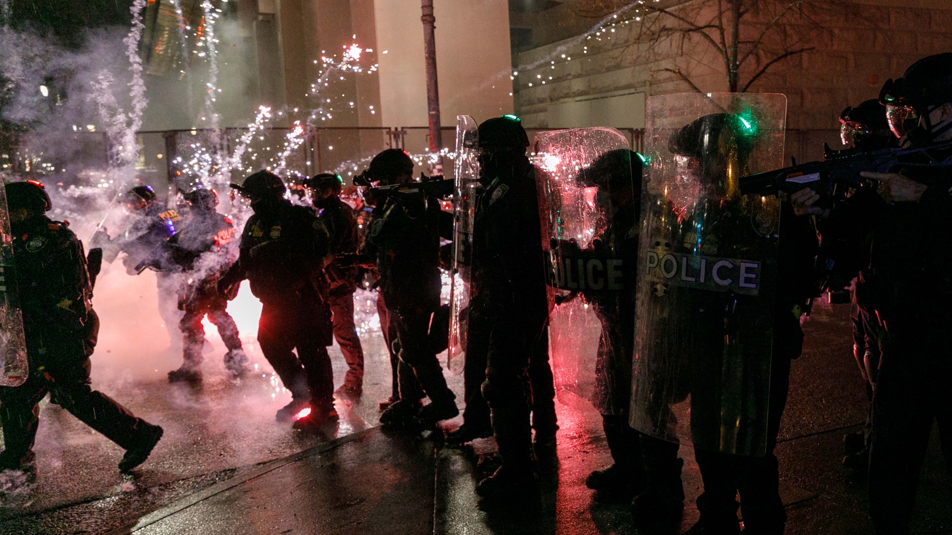 A line of people in police body protection stand in a line with an explosion off to the left of the frame.