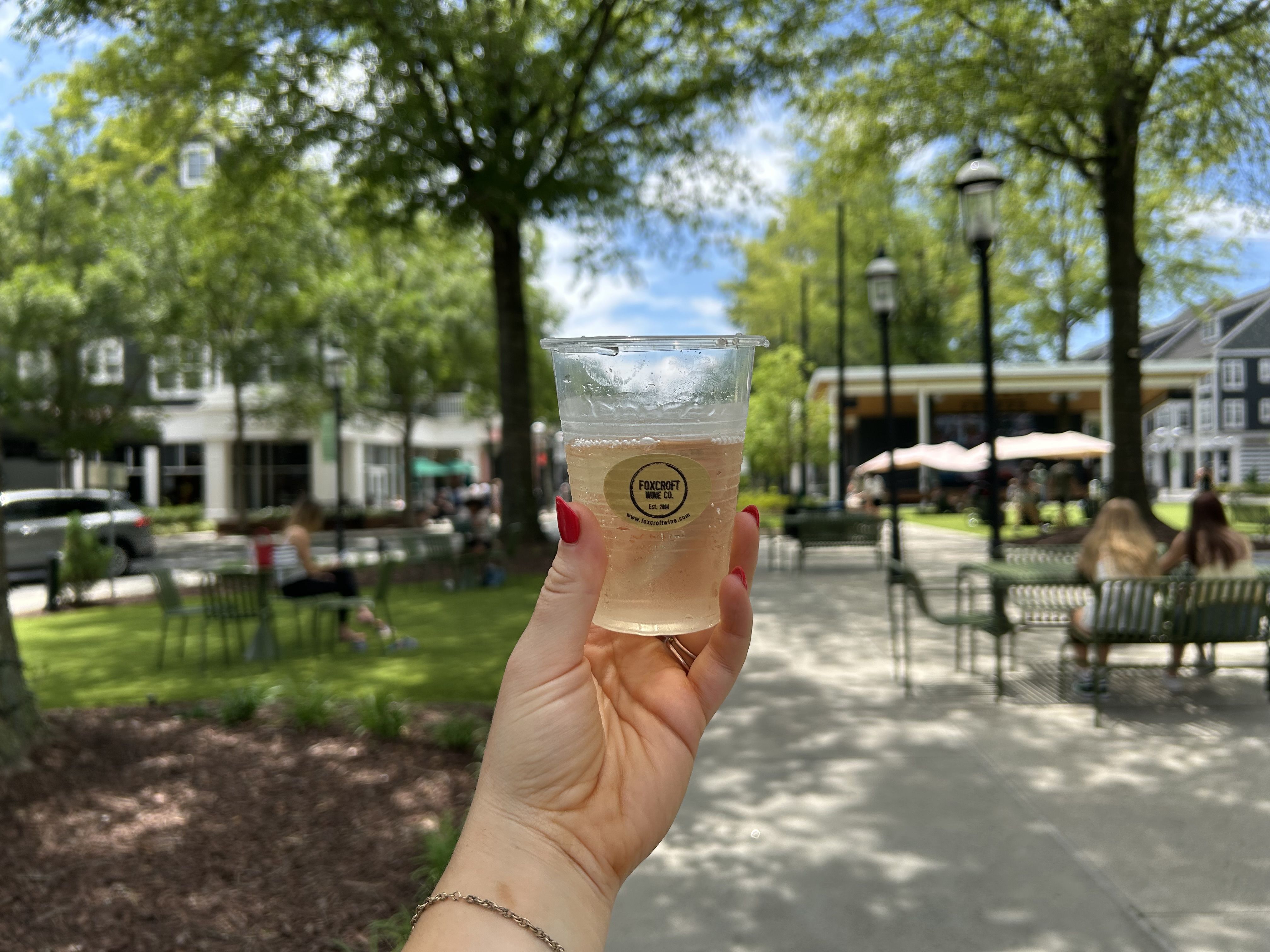 Hand with red nails holding a clear plastic cup of light-colored drink labeled Foxcroft Wine Co. in a sunny park area with green trees, benches, people, and buildings in the background.