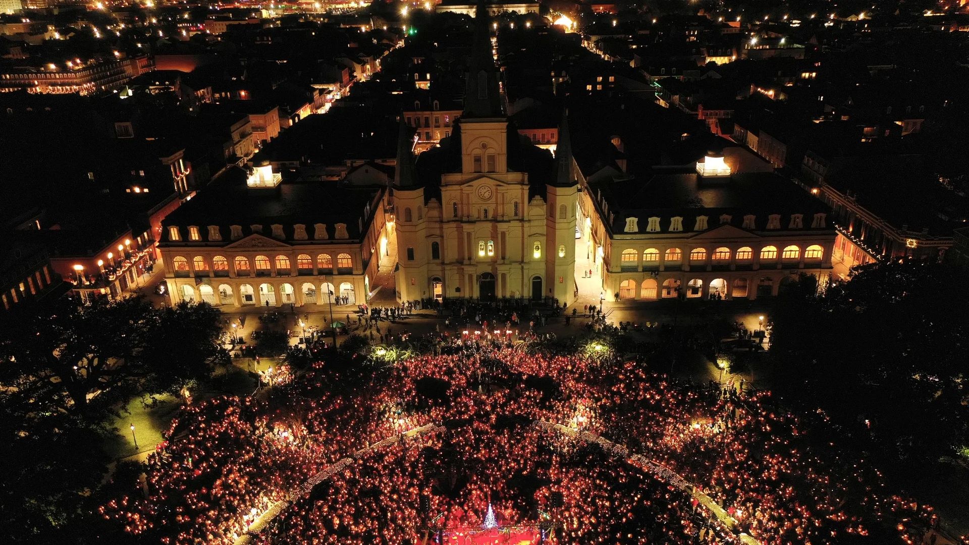 A large crowd holding red lights gathers at night in front of an illuminated historic building with three towers, surrounding a Christmas tree on a stage.