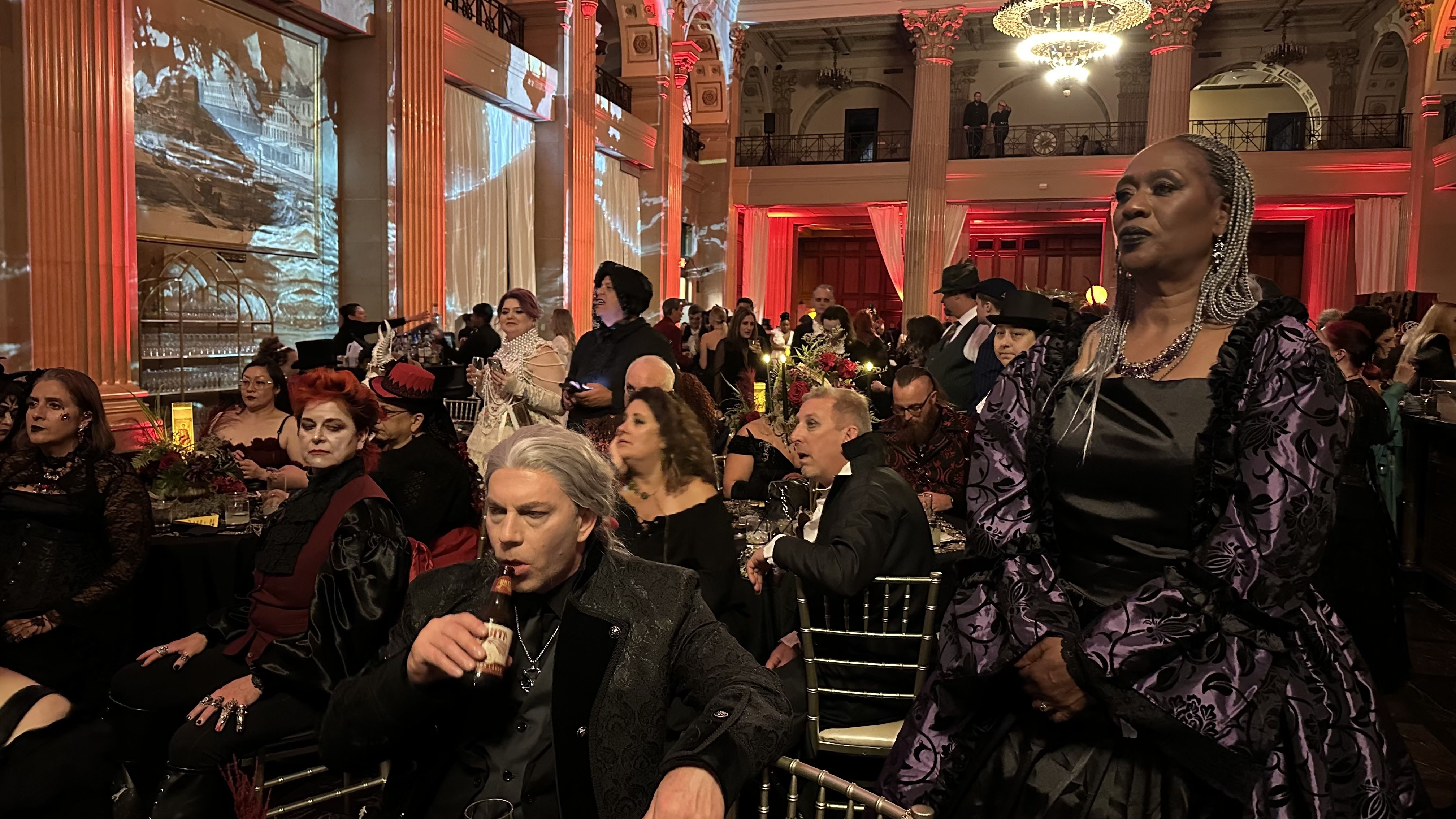 People in gothic and Victorian-style costumes at a formal indoor event with red-lit columns and chandeliers, including a woman in a purple and black dress and a man drinking from a bottle.