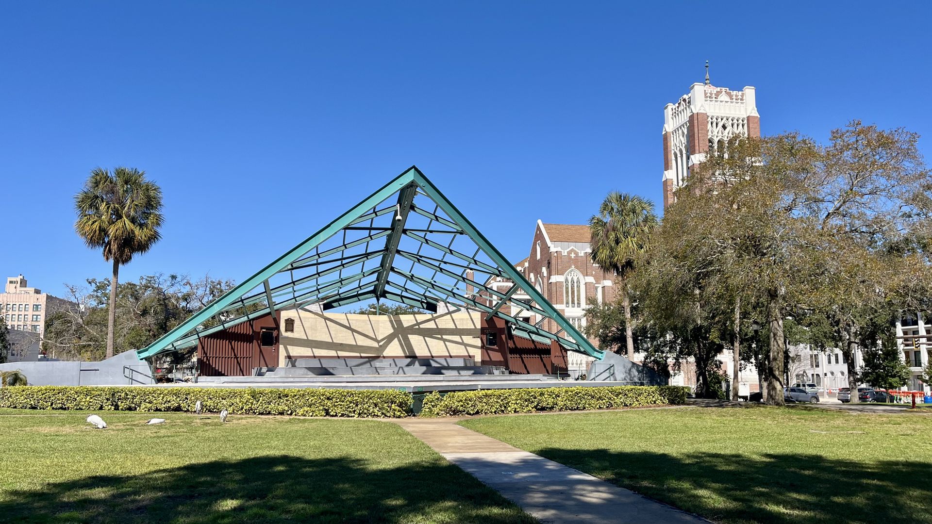 Sunny park scene with a teal-framed glass pyramidal pavilion, a concrete path, and green lawn with hedges. Palm trees line the area; a tall brick building rises behind trees against a clear blue sky.