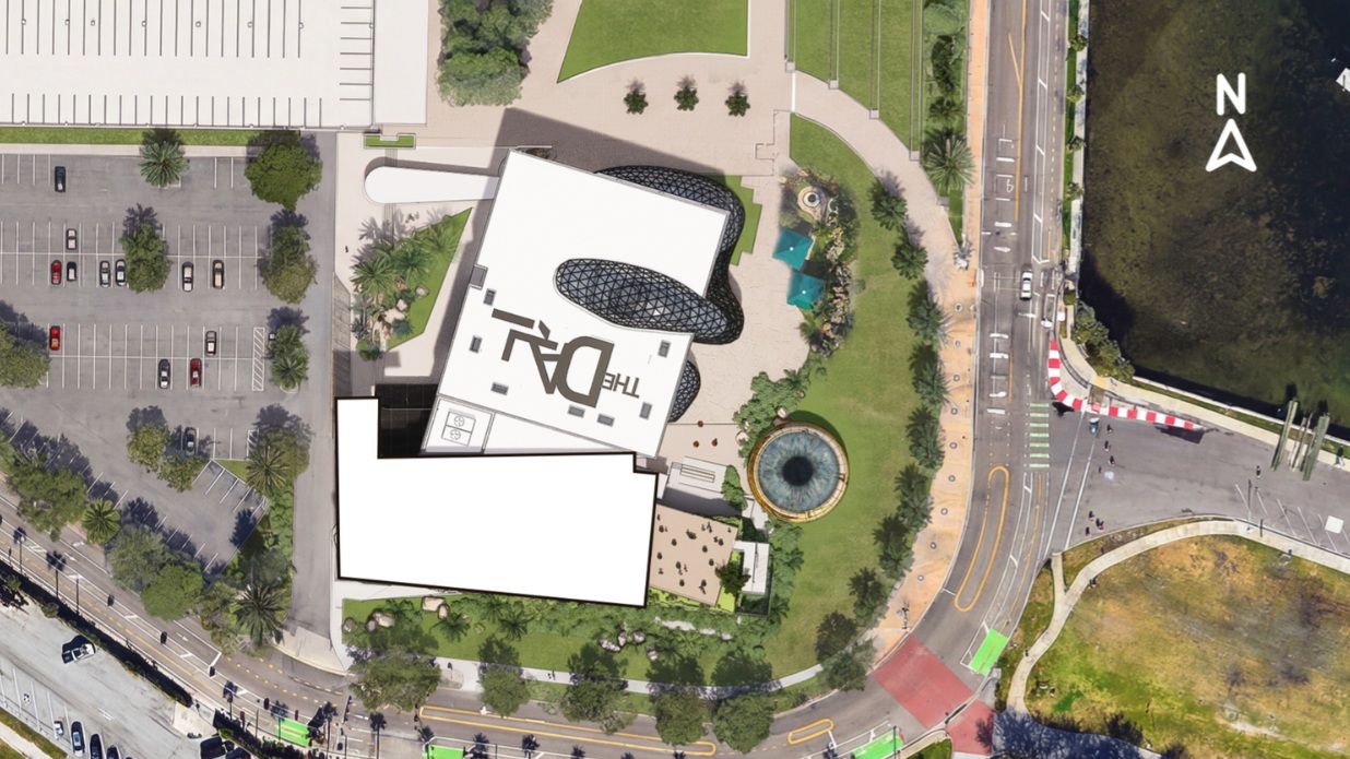 Aerial view of The Dalí Museum with white roofs and black glass domes, surrounded by green lawns, a circular fountain, parking lots, roads, and a water body on the right.