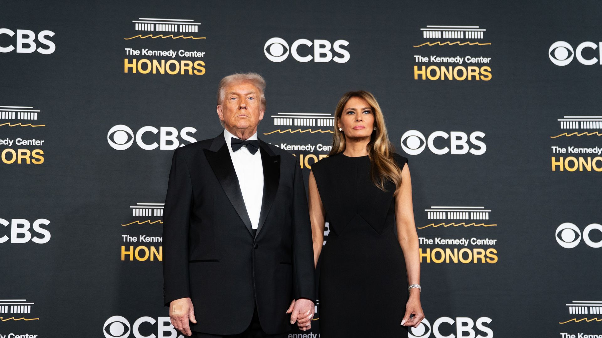 President Trump, wearing a tuxedo, holds hands with Melania Trump, who is wearing a black dress, at a step-and-repeat for the Kennedy Center Honors.