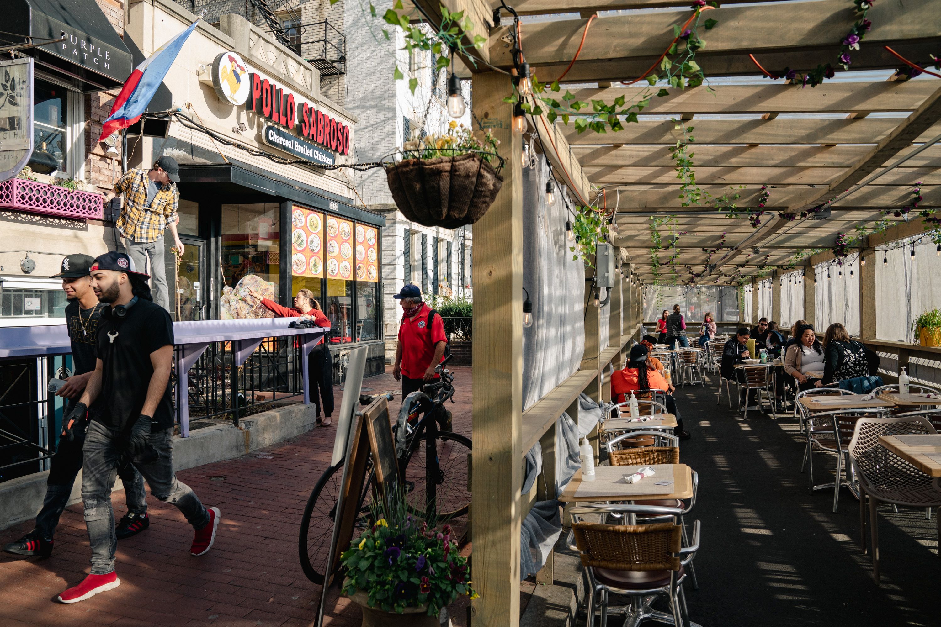 Outdoor street scene with the Pollo Sabroso restaurant sign and a flag; people walk by and dine at metal tables under a wooden trellis with hanging lights and greenery.