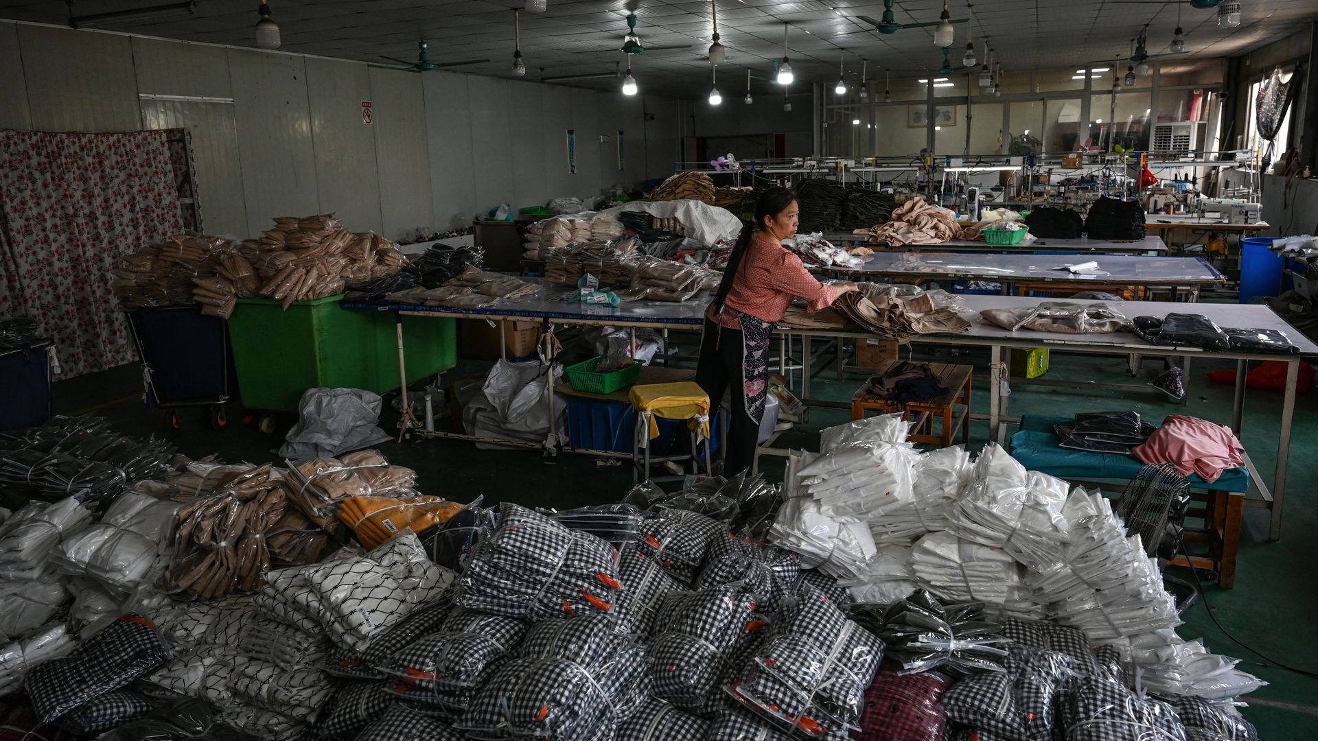 Woman organizing piles of folded clothing in a large, dimly lit textile workshop filled with stacks of packaged clothes in various colors, sewing tables, and ceiling fans.