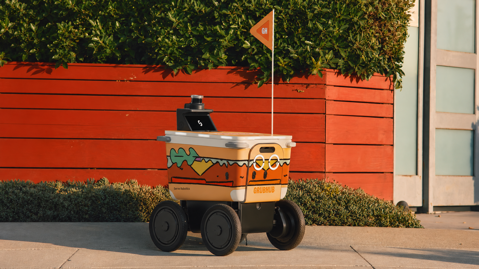 A four-wheeled delivery robot with an orange Grubhub container sits on a sidewalk, a GH flag on a pole, before a red wooden fence and green hedges beside a glass building.