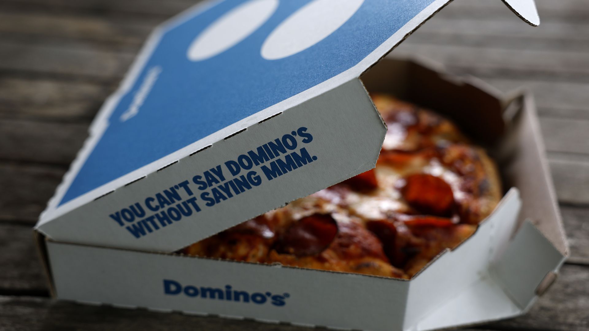 Partially open Domino's pizza box on wooden surface showing pepperoni pizza inside. Text on box reads, "You can't say Domino's without saying mmm."