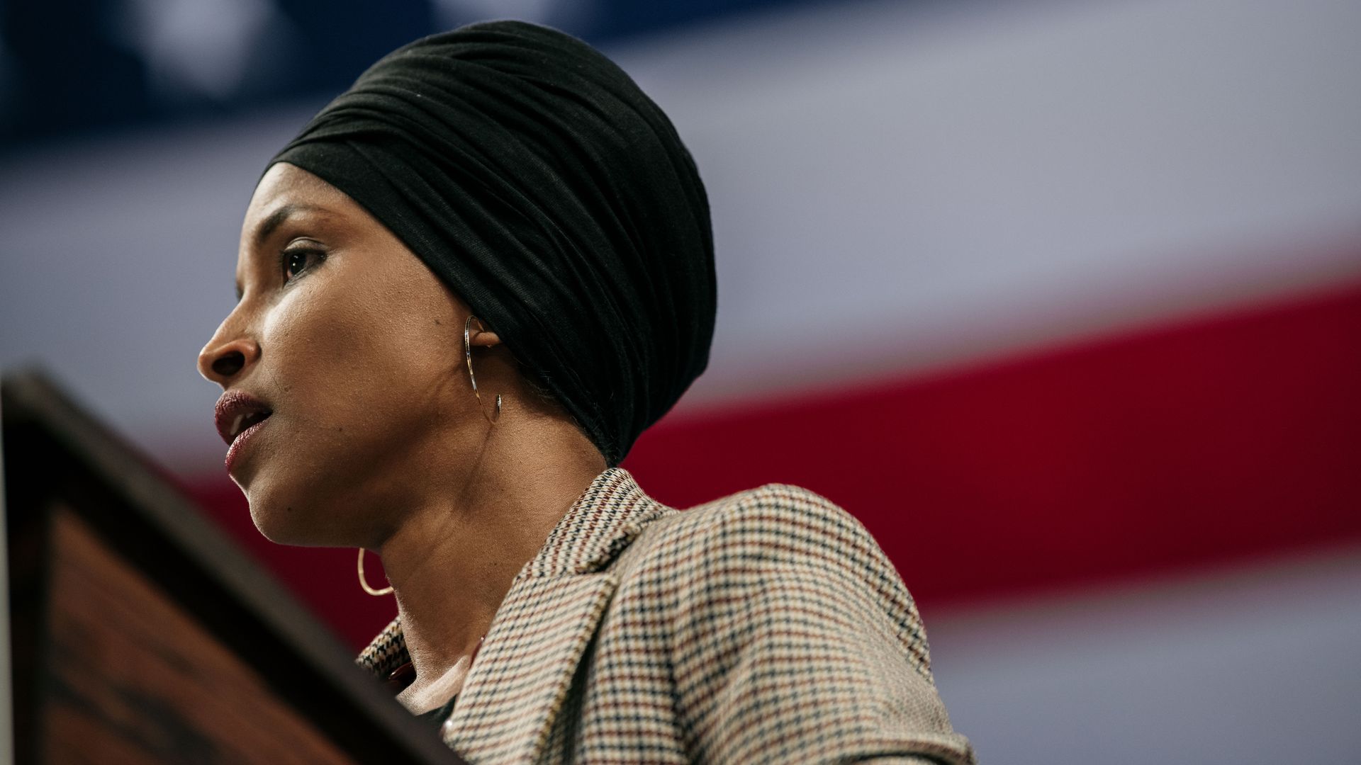 In this image, Omar stands in front of a podium with a large American flag behind her.