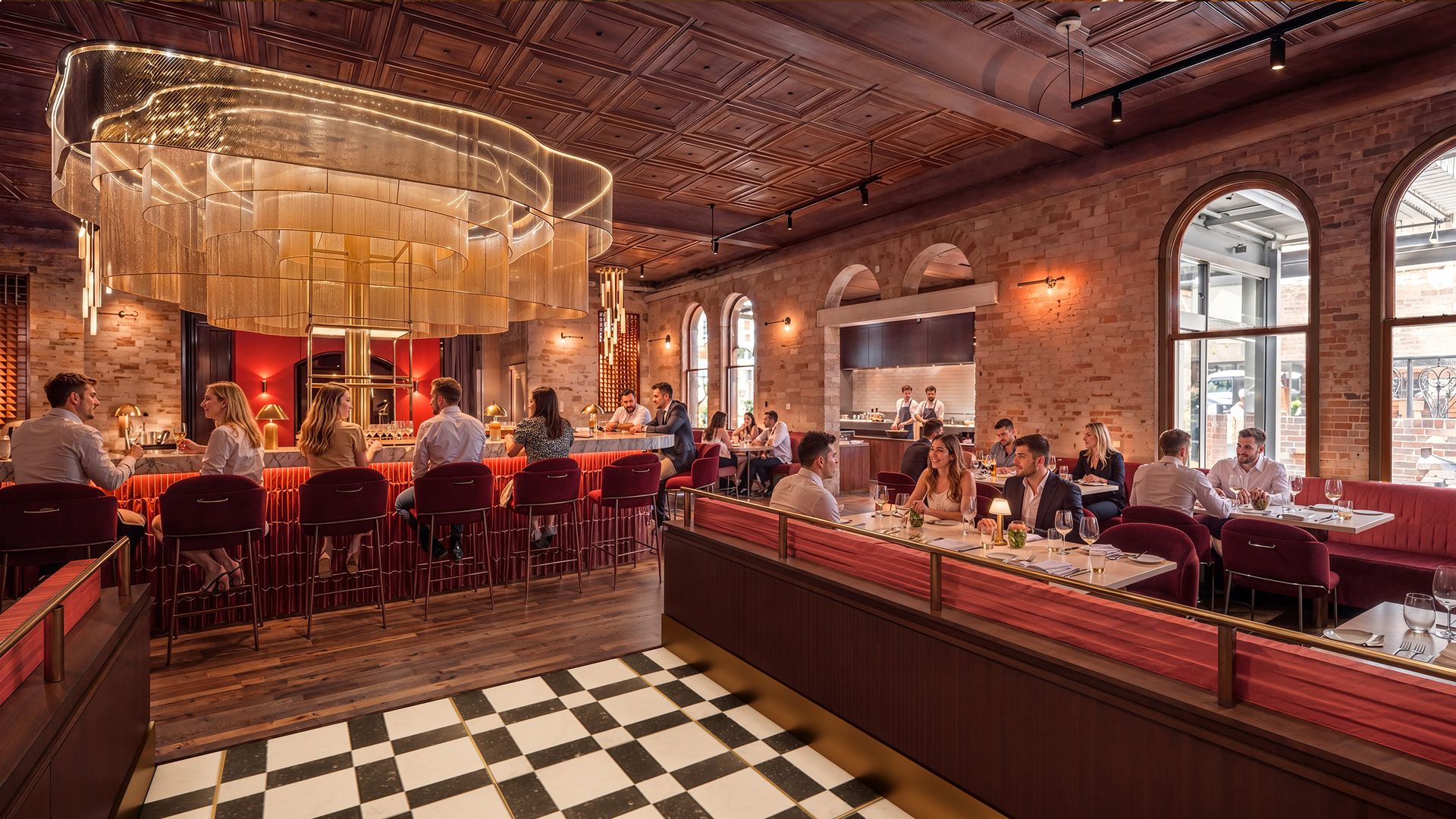 Upscale restaurant interior with a large gold circular chandelier, brick walls, arched windows, red velvet chairs, groups of diners at tables, and a black-and-white checkered floor.