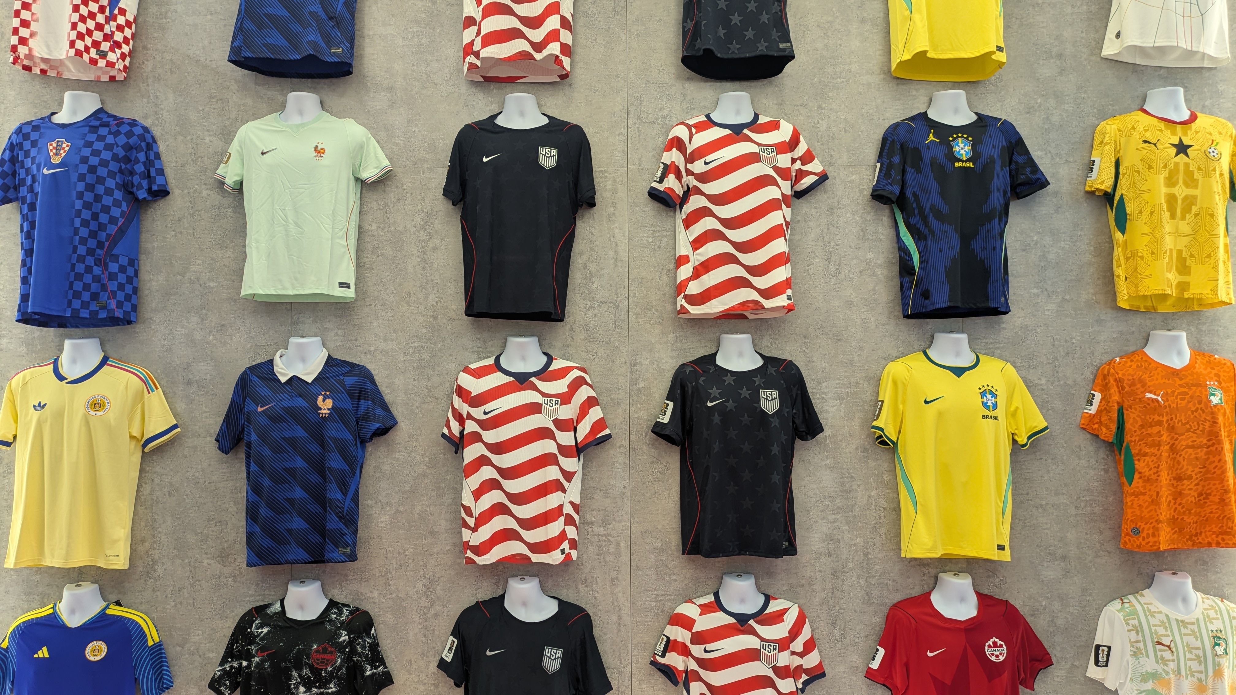 Rows of colorful soccer jerseys hung on a gray wall, featuring USA black and striped kits, mint France, Brazil yellow, Colombia yellow, Croatia red-white checks, plus blue and navy designs.