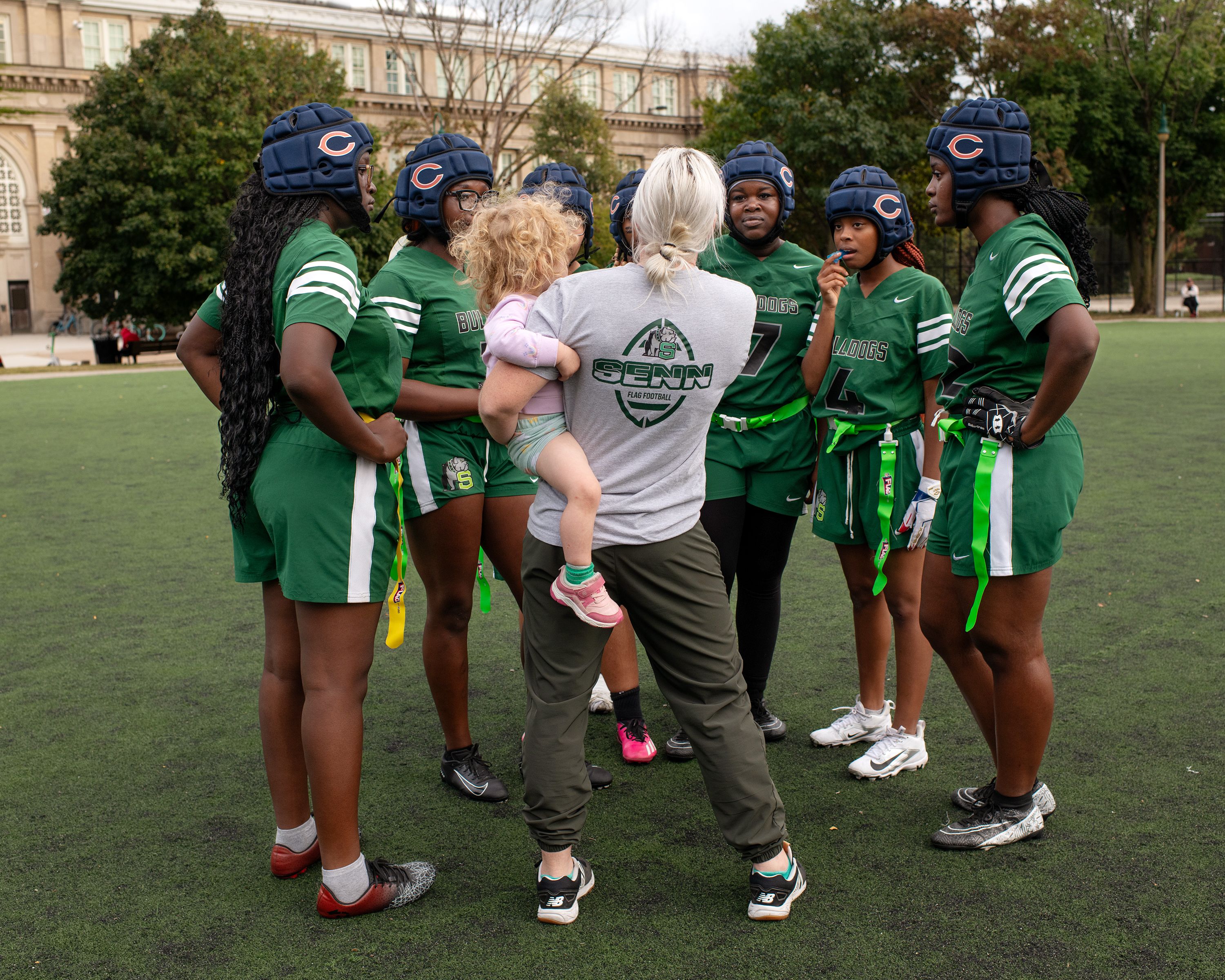 Coach holding child talks to female flag football players in green uniforms and navy helmets on a field, forming a huddle during practice or game break.
