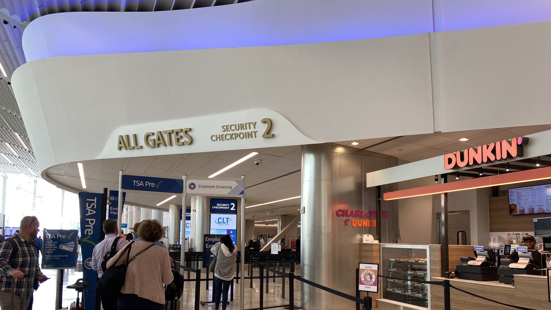 Travelers wait in line at airport Security Checkpoint 2 near TSA Pre area, with a Dunkin' coffee shop on the right and signage for all gates above; ceiling lit with blue lighting.