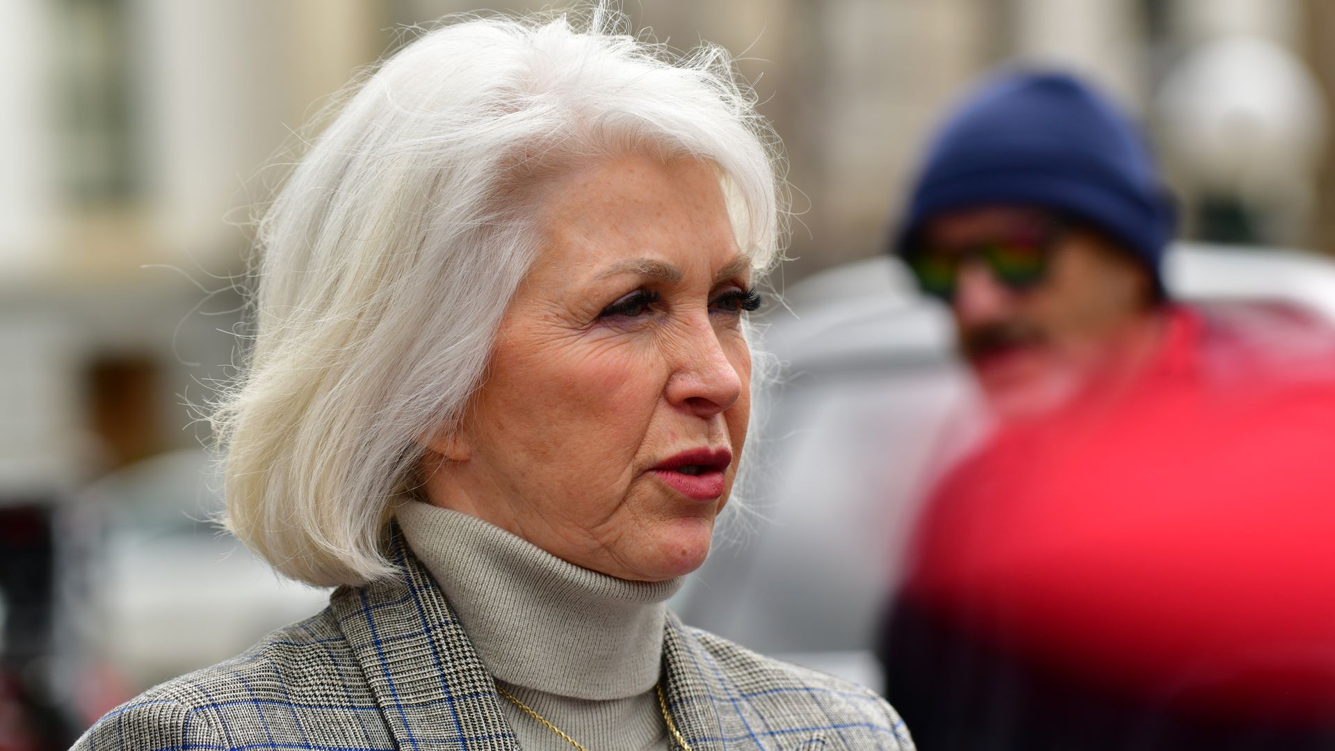 Tina Peters wearing a beige turtleneck and plaid blazer, outdoors with blurred people and vehicles in the background.