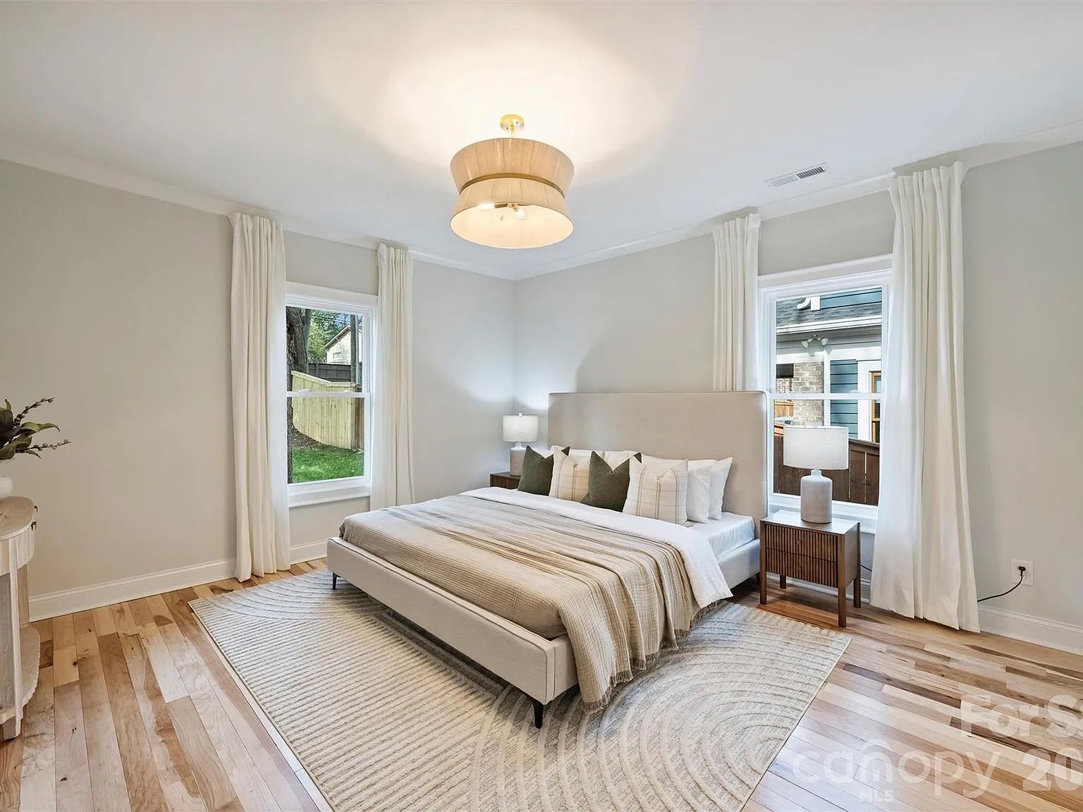 A bright bedroom with a large upholstered bed, beige headboard, two wooden nightstands with white lamps, light hardwood floors, a neutral striped rug, and tall white curtains framing two windows.