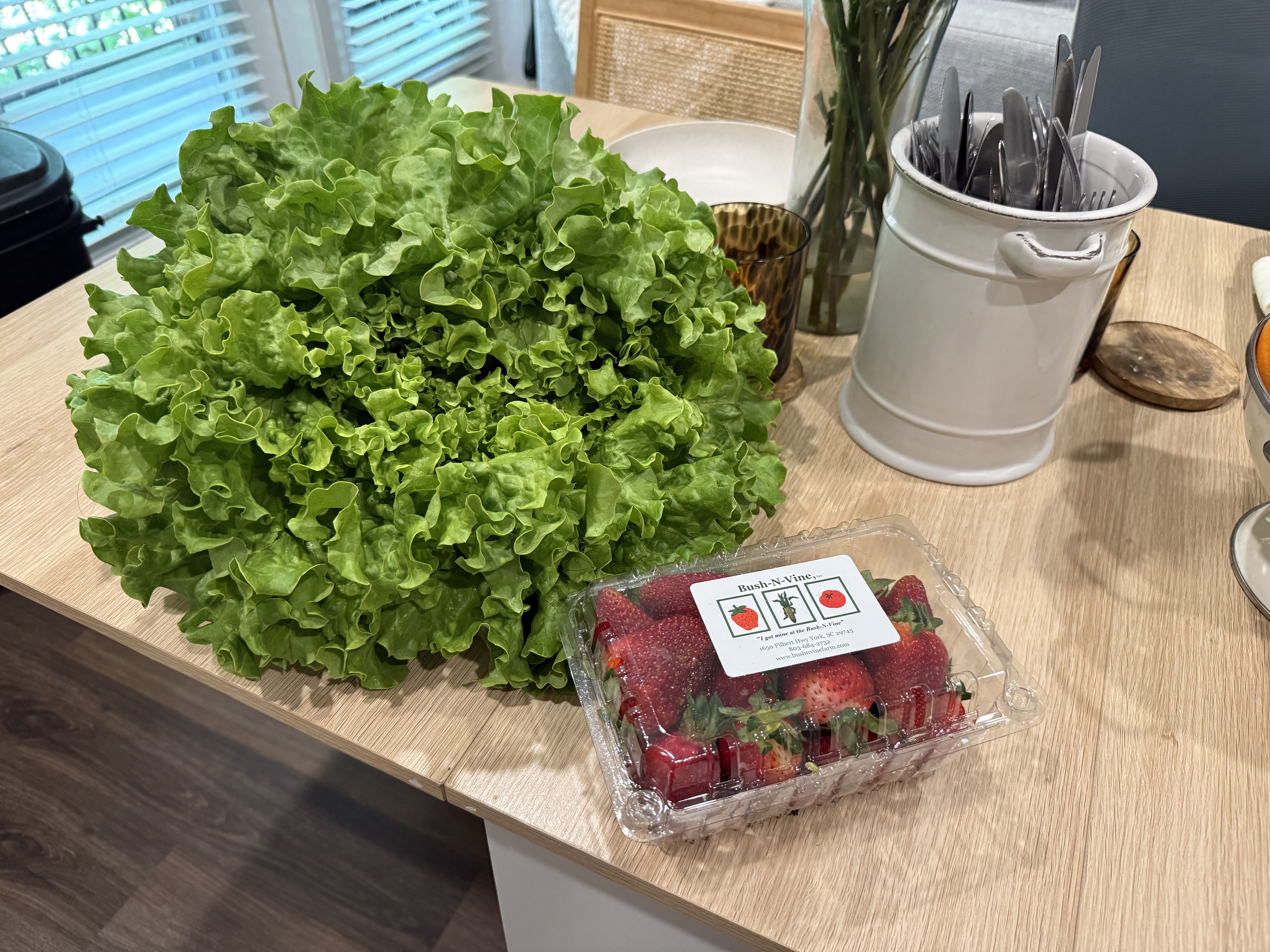 Large head of green lettuce on a wooden kitchen table beside a white utensil holder filled with forks and knives; a clear clamshell of red strawberries labeled Bush-N-Vine sits nearby.