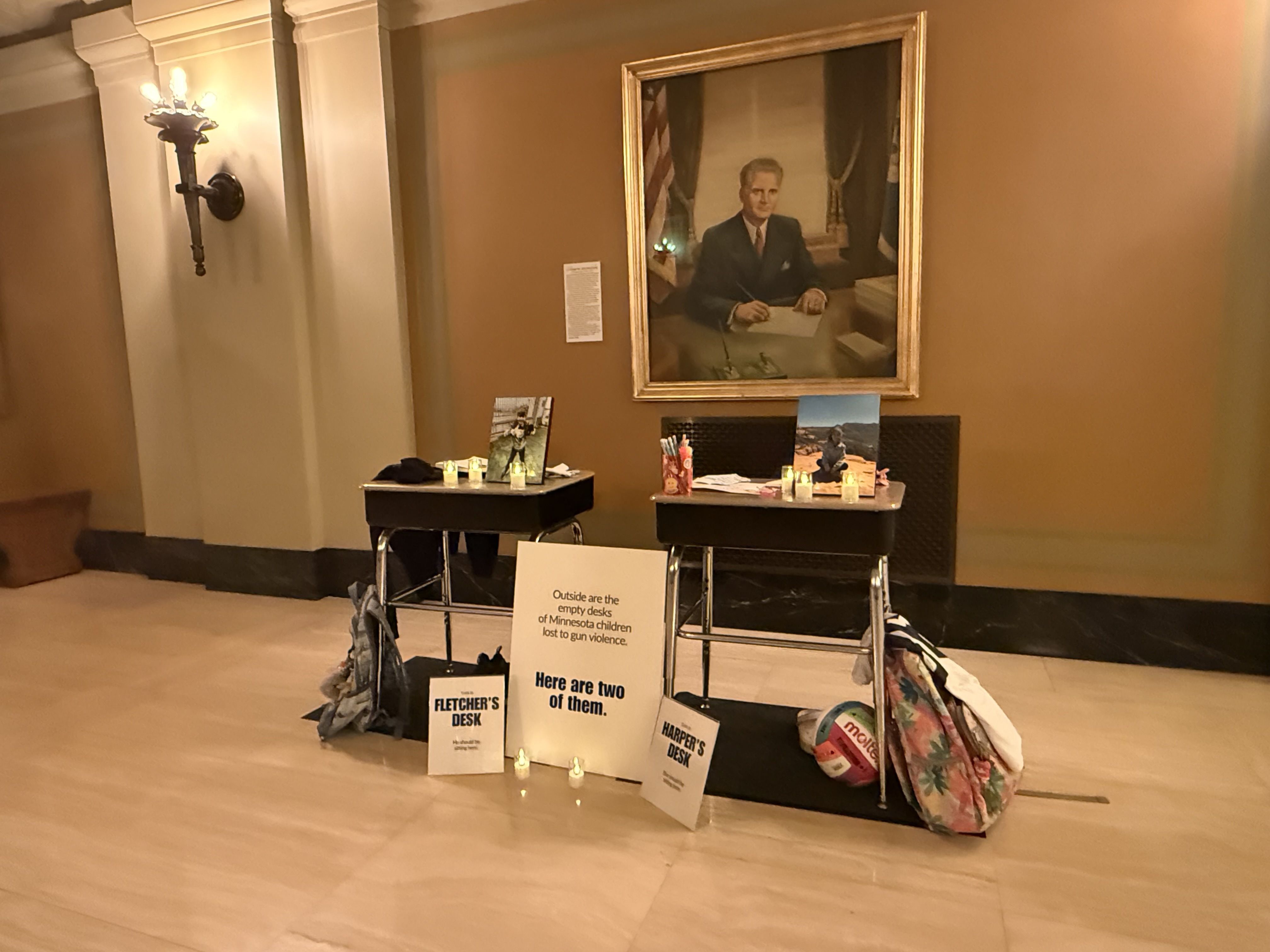 Two school desks displayed with candles, photos, backpacks, and signs in a room with beige walls and a portrait. Signs highlight Minnesota children lost to gun violence.