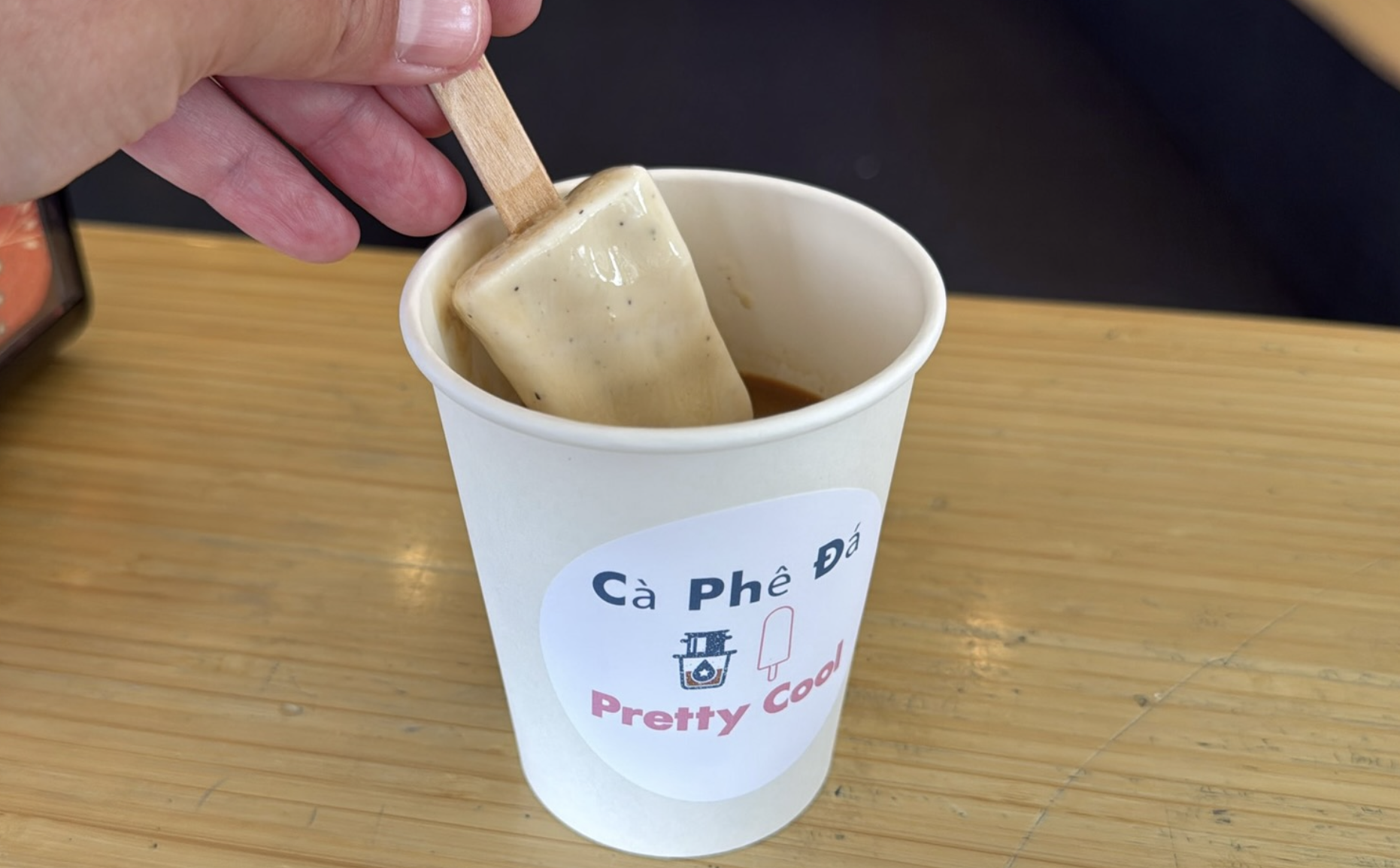 Close-up of a hand pulling a speckled beige coffee ice pop from a white paper cup labeled "Pretty Cool" on a wooden table.