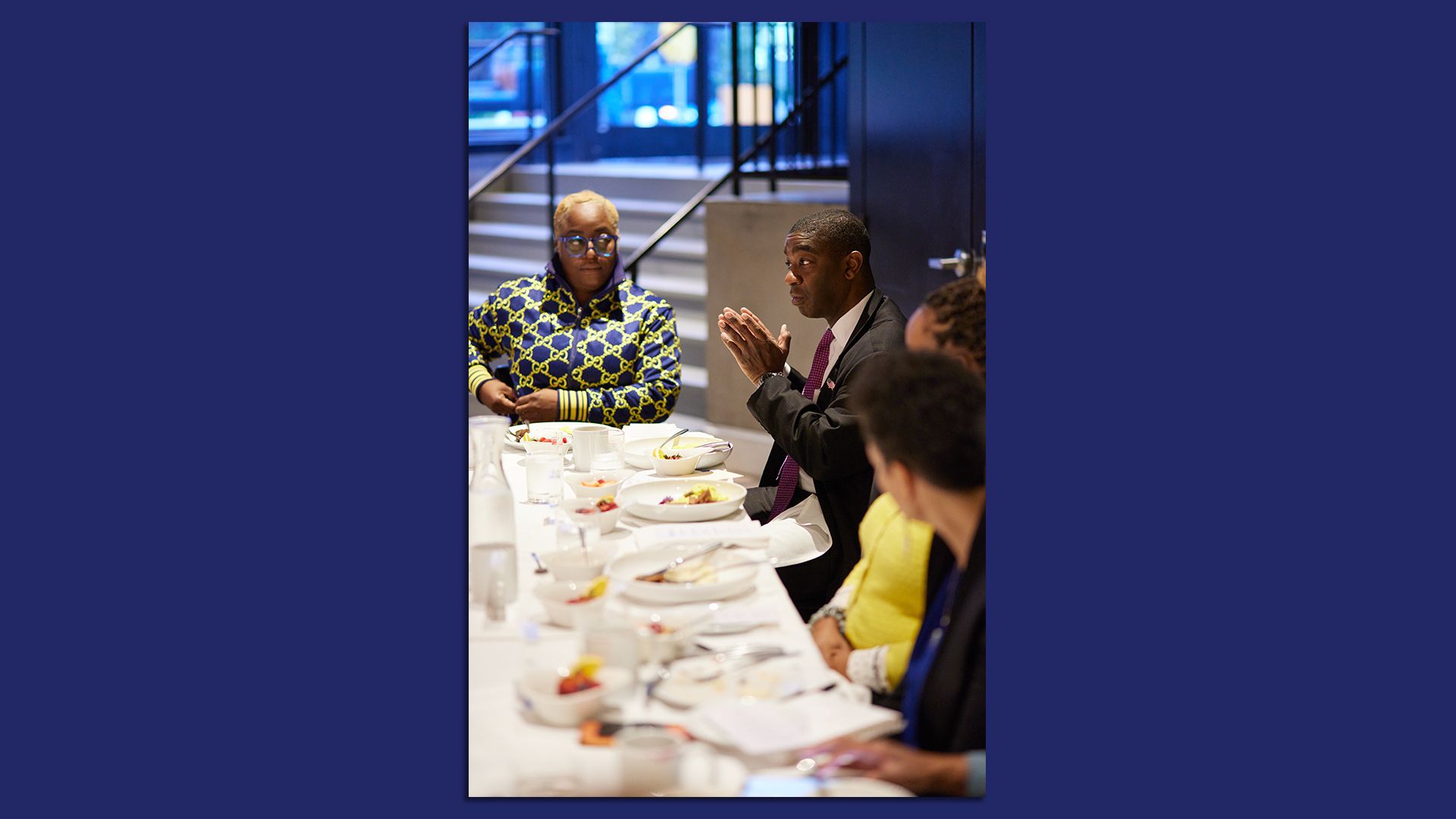 A man speaks to a group of people sitting around a large square table with a white tablecloth.