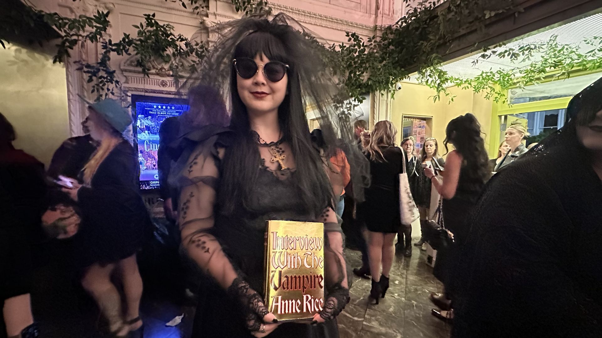 Person dressed in black Gothic attire with veil and sunglasses holds a gold book titled "Interview With The Vampire Anne Rice" at a crowded event with people and greenery around.